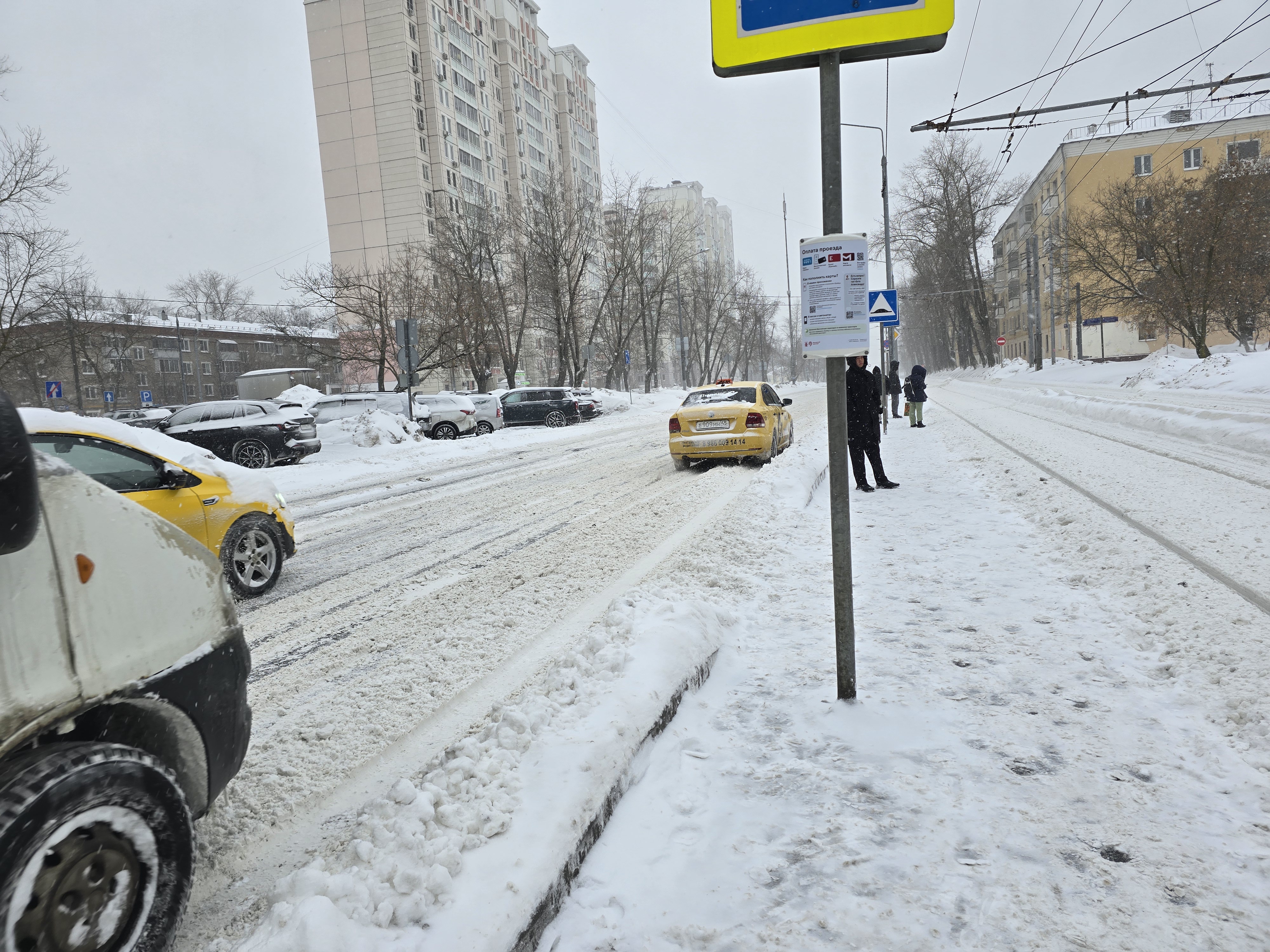 Участок городской дороги без пешеходного перехода, покрытый снегом. Видны припаркованные автомобили и люди на тротуаре.