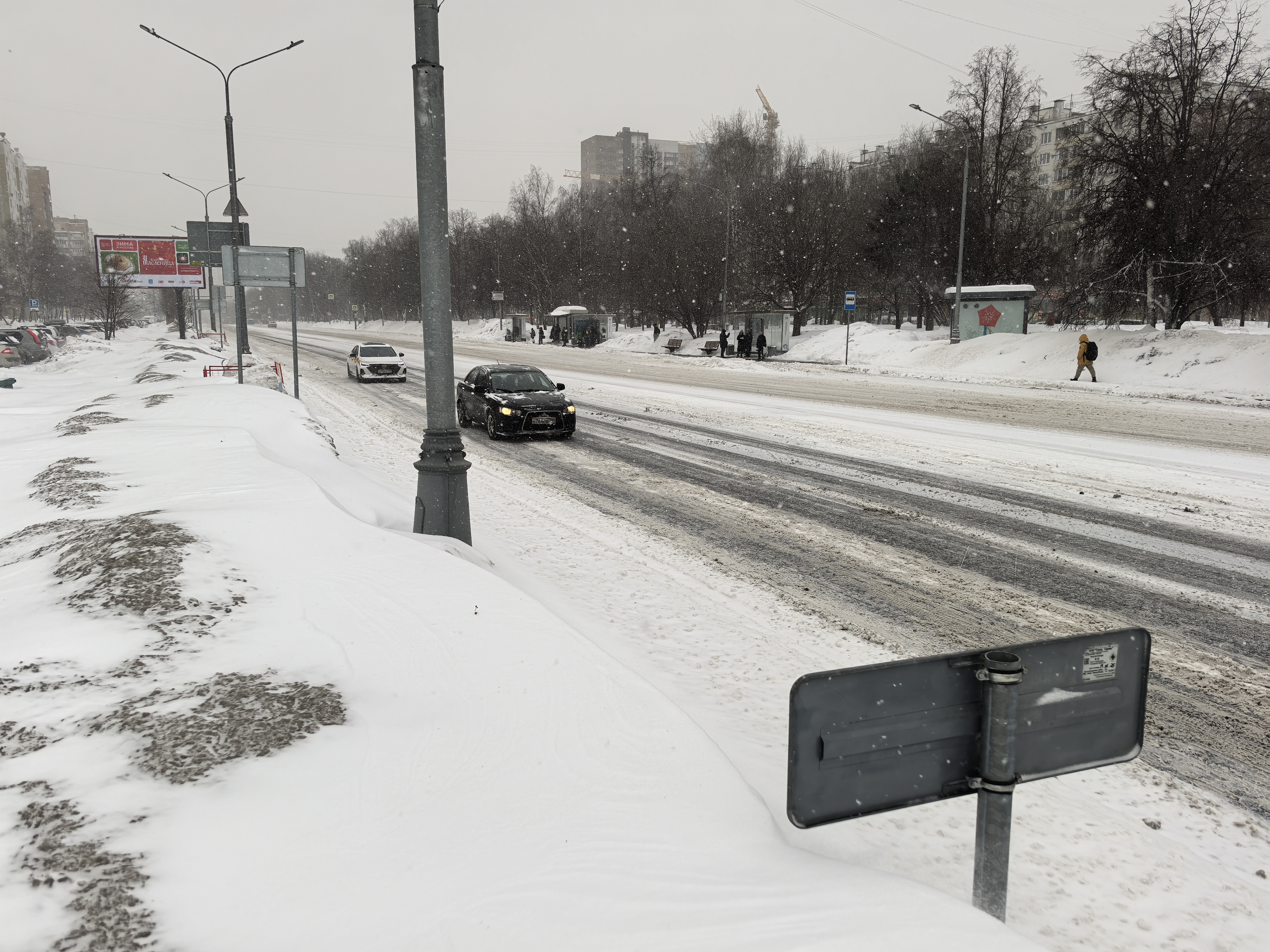 Широкая городская дорога в зимний период, покрытая снегом и слякотью. Пешеходный переход отсутствует, движение осуществляется по тротуару и обочине.