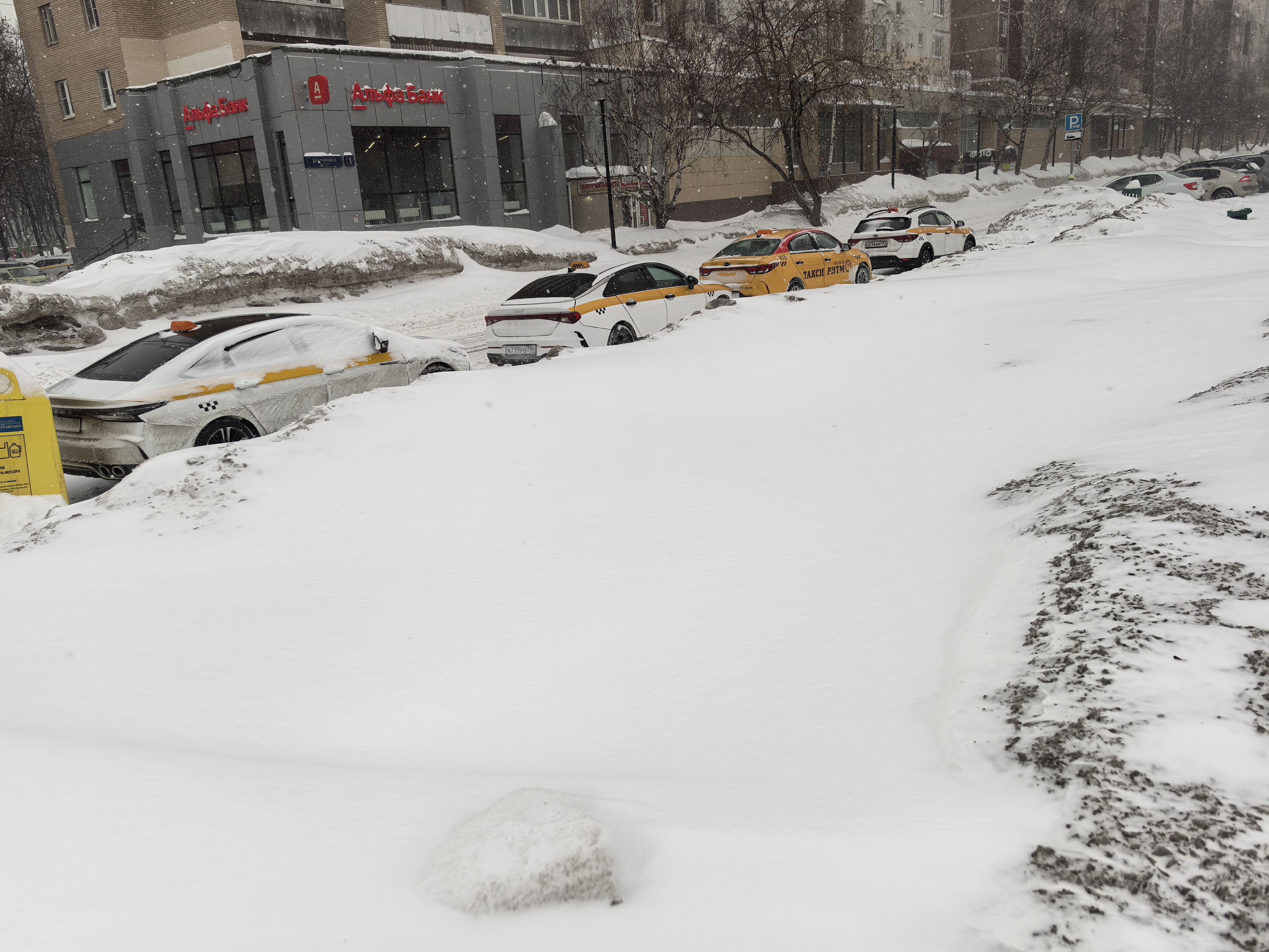Участок проезжей части в зимний период, полностью покрытый снегом. Пешеходный переход отсутствует. Видны припаркованные такси вдоль дороги и здание Альфа-Банка на заднем плане.