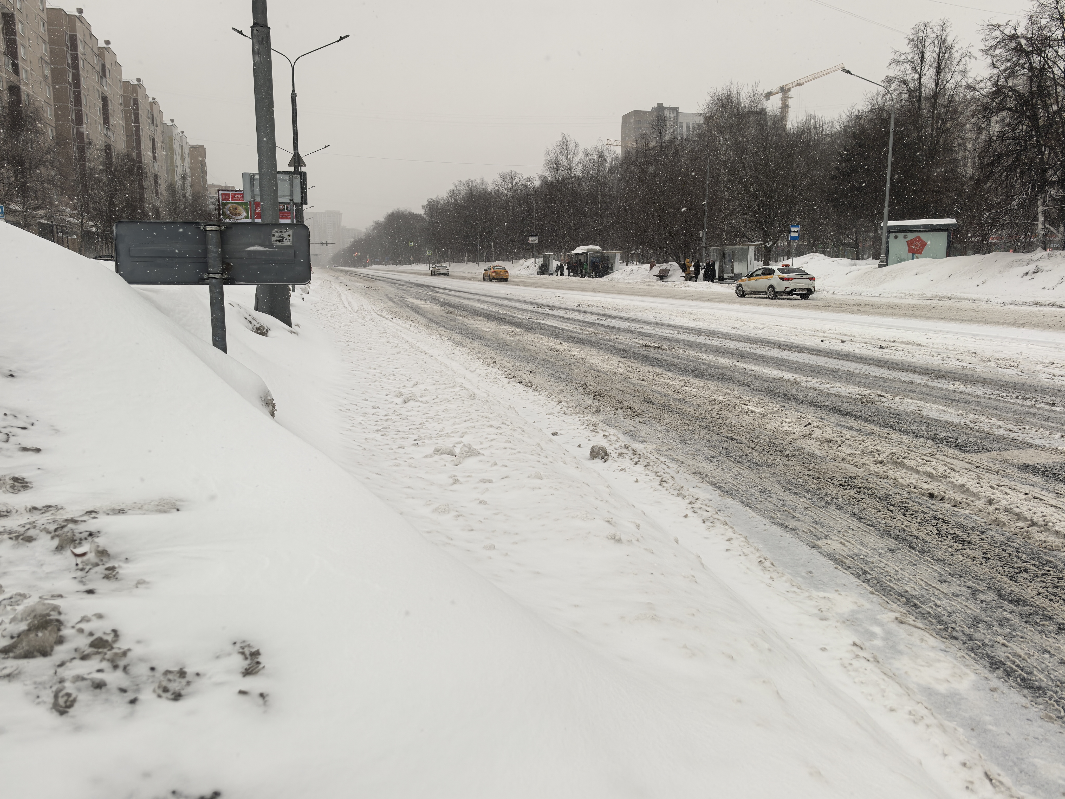 Широкая городская магистраль в зимний период. Дорога покрыта снегом и слякотью, тротуары и обочины занесены снегом. Пешеходный переход на данном участке визуально отсутствует, движение осуществляется по проезжей части.