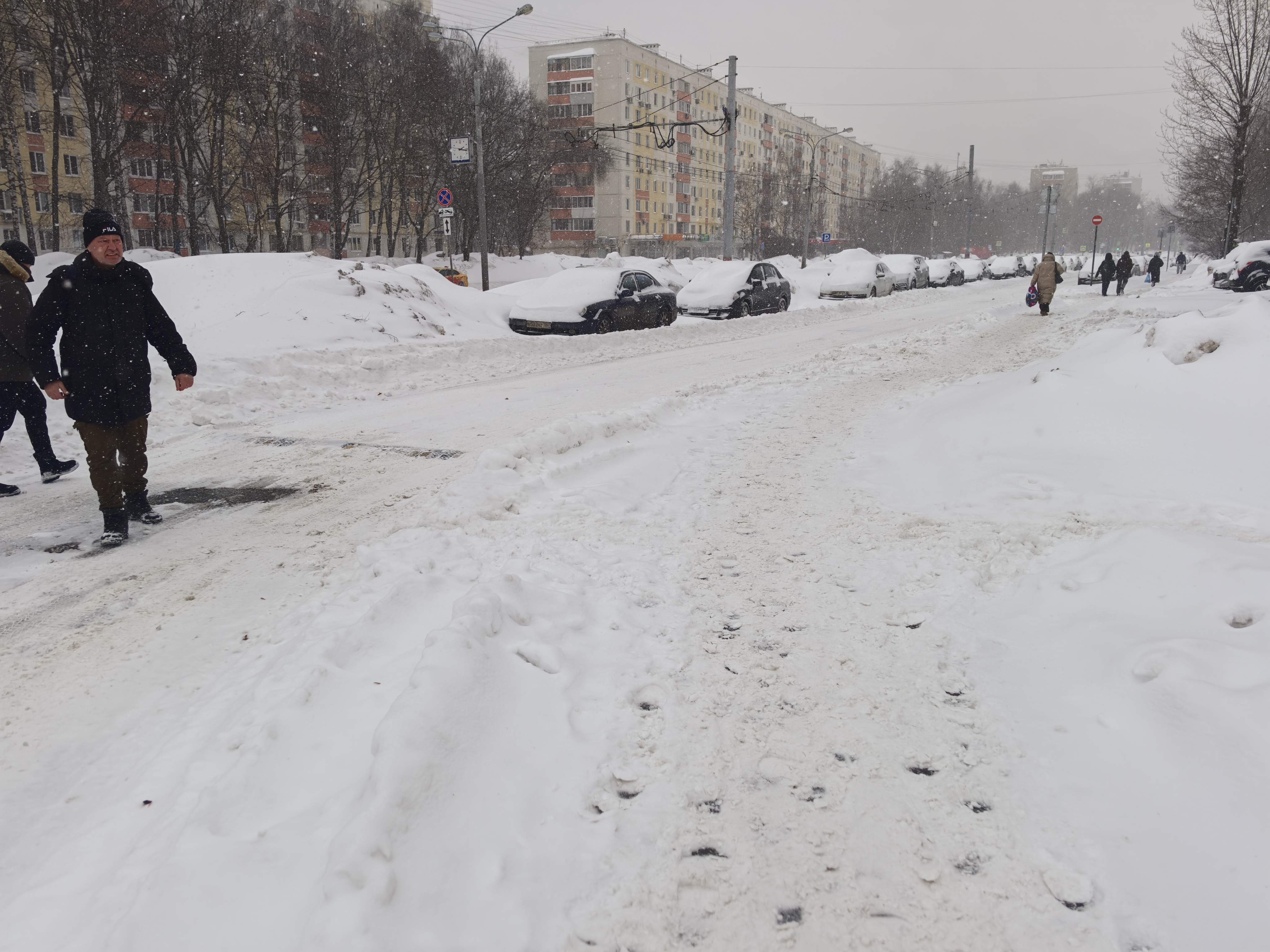 Участок городской дороги в зимний период, покрытый снегом и наледью. Пешеходный переход отсутствует, движение пешеходов осуществляется по проезжей части или неочищенным обочинам. Видны припаркованные автомобили, полностью покрытые снегом.