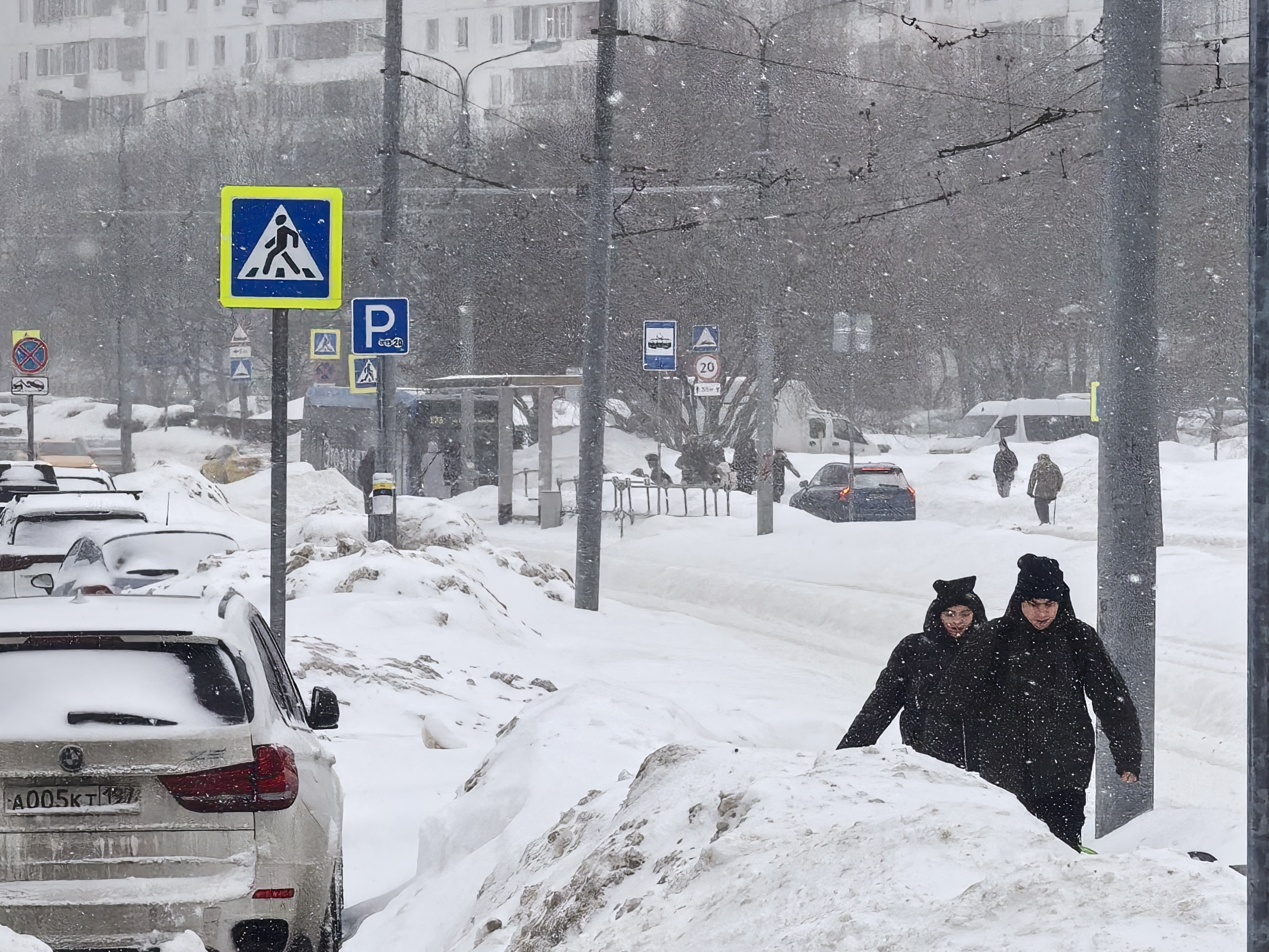 Городская улица в зимний период с пешеходным переходом, обозначенным знаком и разметкой, покрытой снегом. Видны припаркованные автомобили и движение транспорта.