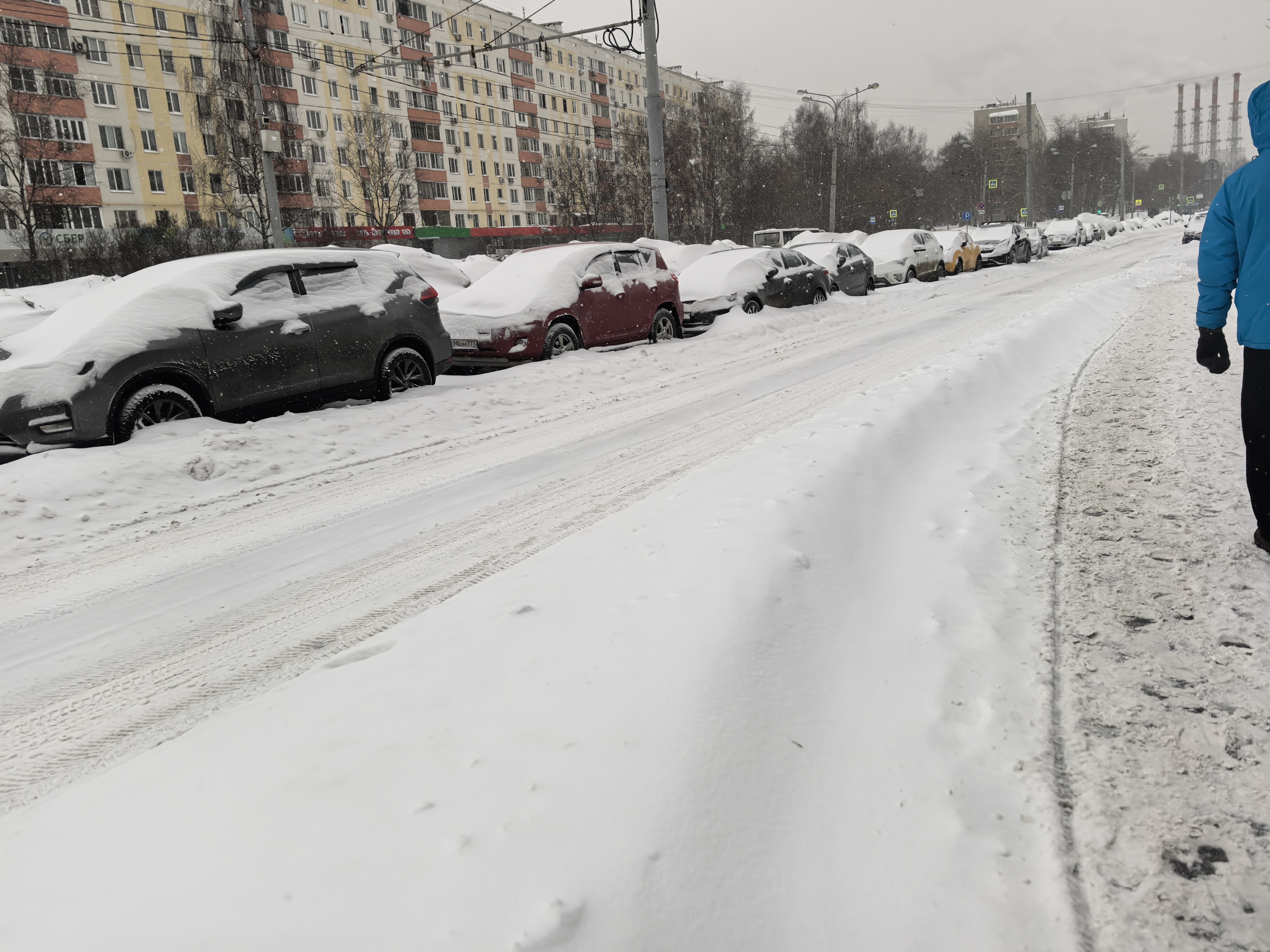 Городская улица в зимний период, покрытая снегом. Парковка автомобилей вдоль бордюра, тротуар частично очищен, но пешеходный переход в кадре отсутствует.