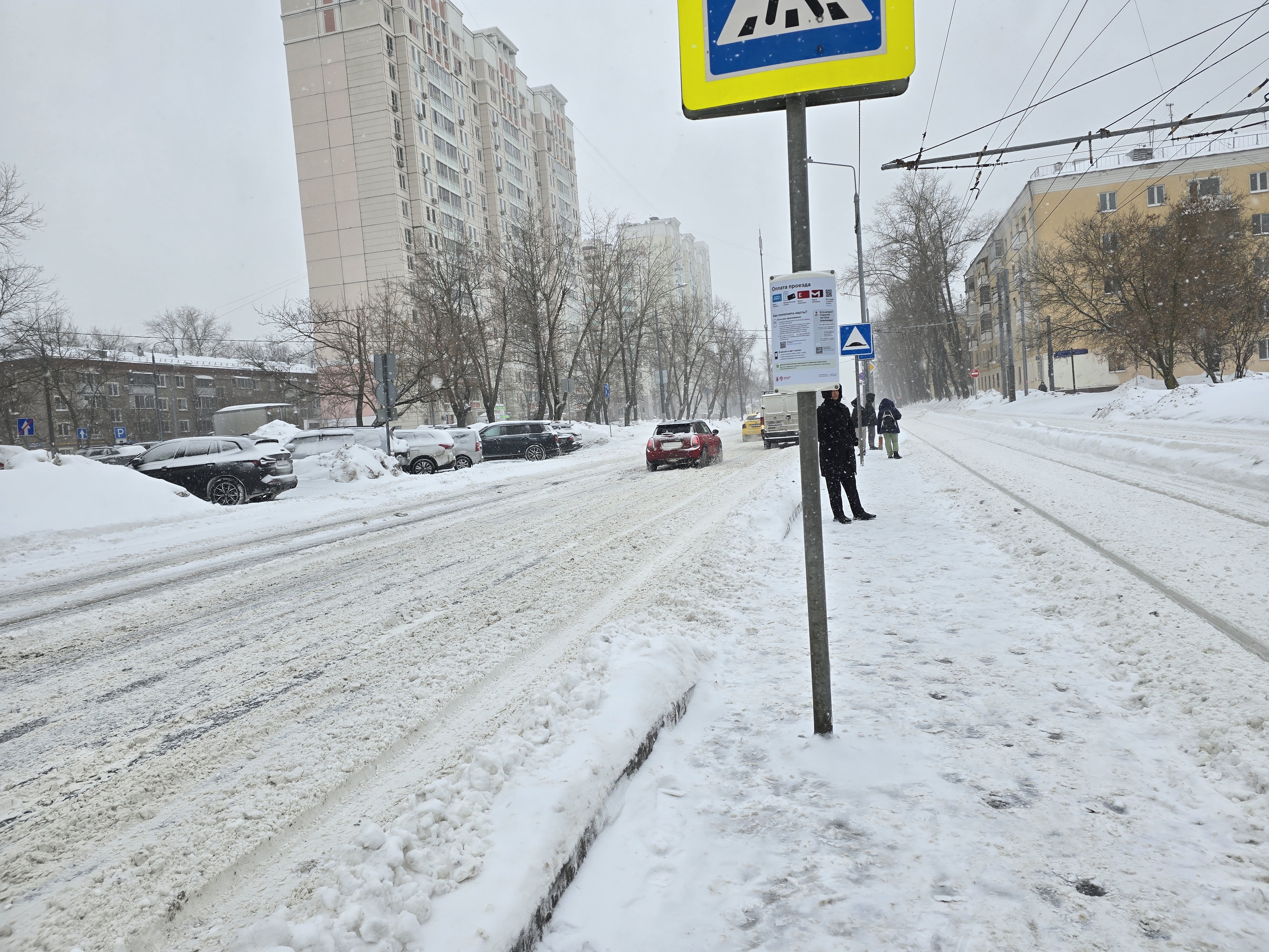 Городская улица с пешеходным переходом в зимних условиях. На проезжей части видна разметка, но она частично скрыта снегом и грязью. Пешеходы стоят на тротуаре, ожидая перехода.