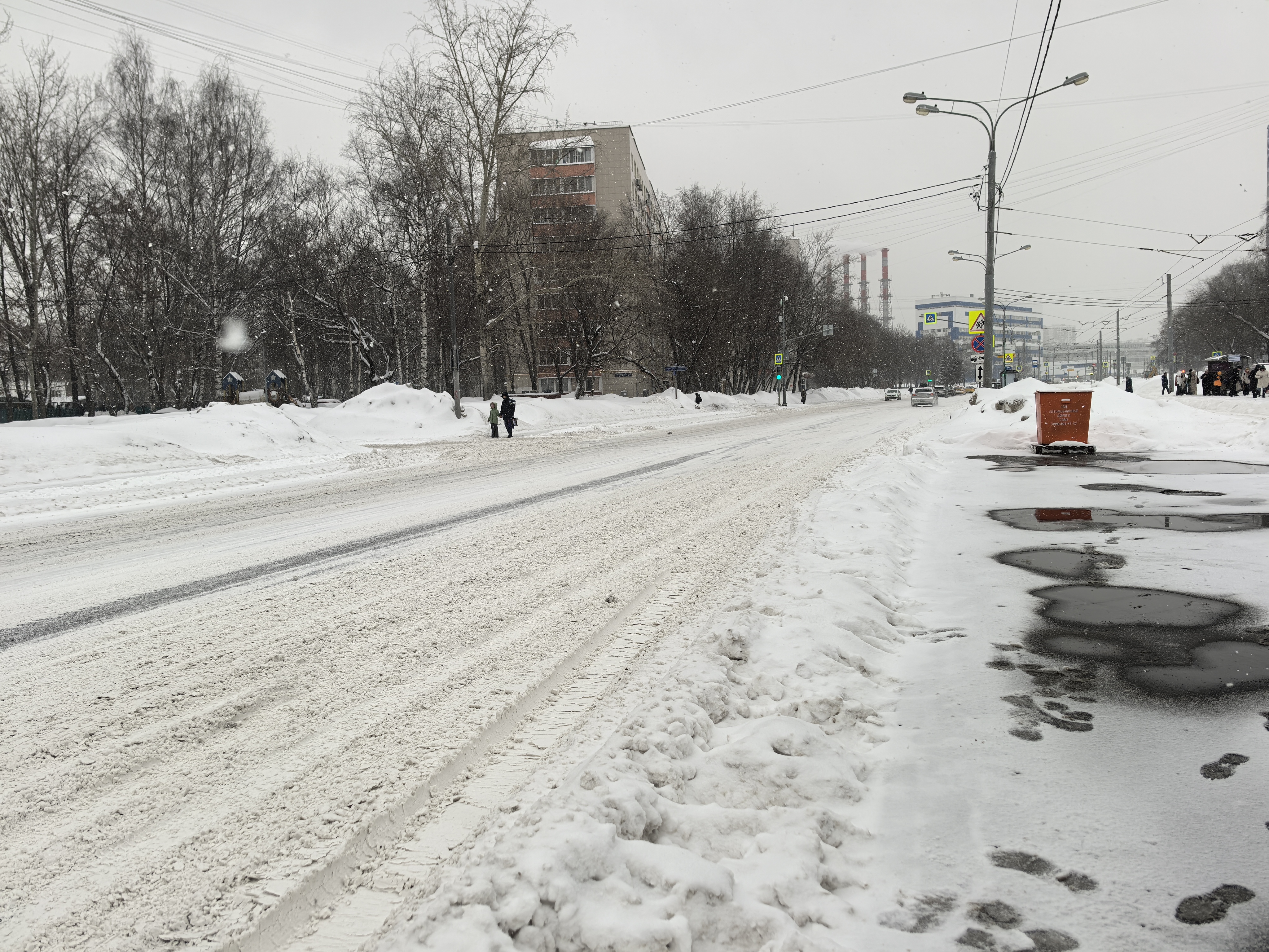 Проезжая часть без пешеходного перехода. Видна широкая дорога, покрытая снегом и слякотью. Тротуары также засыпаны снегом, видны следы пешеходов. На заднем плане видны здания и промышленные трубы.