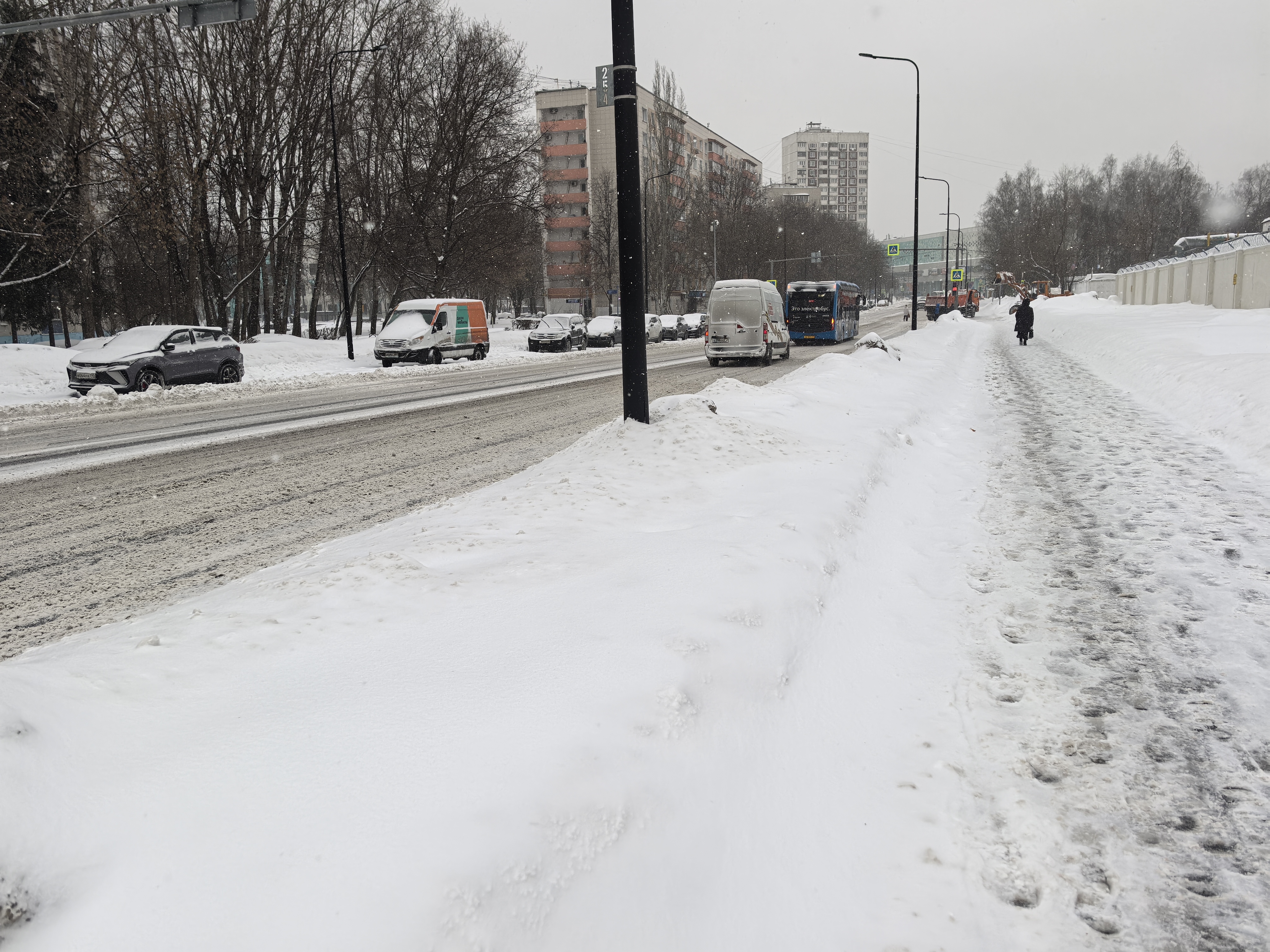 Городская улица в зимний период. Видна проезжая часть с движущимся транспортом (автобус, фургон) и припаркованными машинами. Справа — тротуар, покрытый снегом, с пешеходом. Пешеходный переход в кадре отсутствует, хотя видны знаки вдалеке.