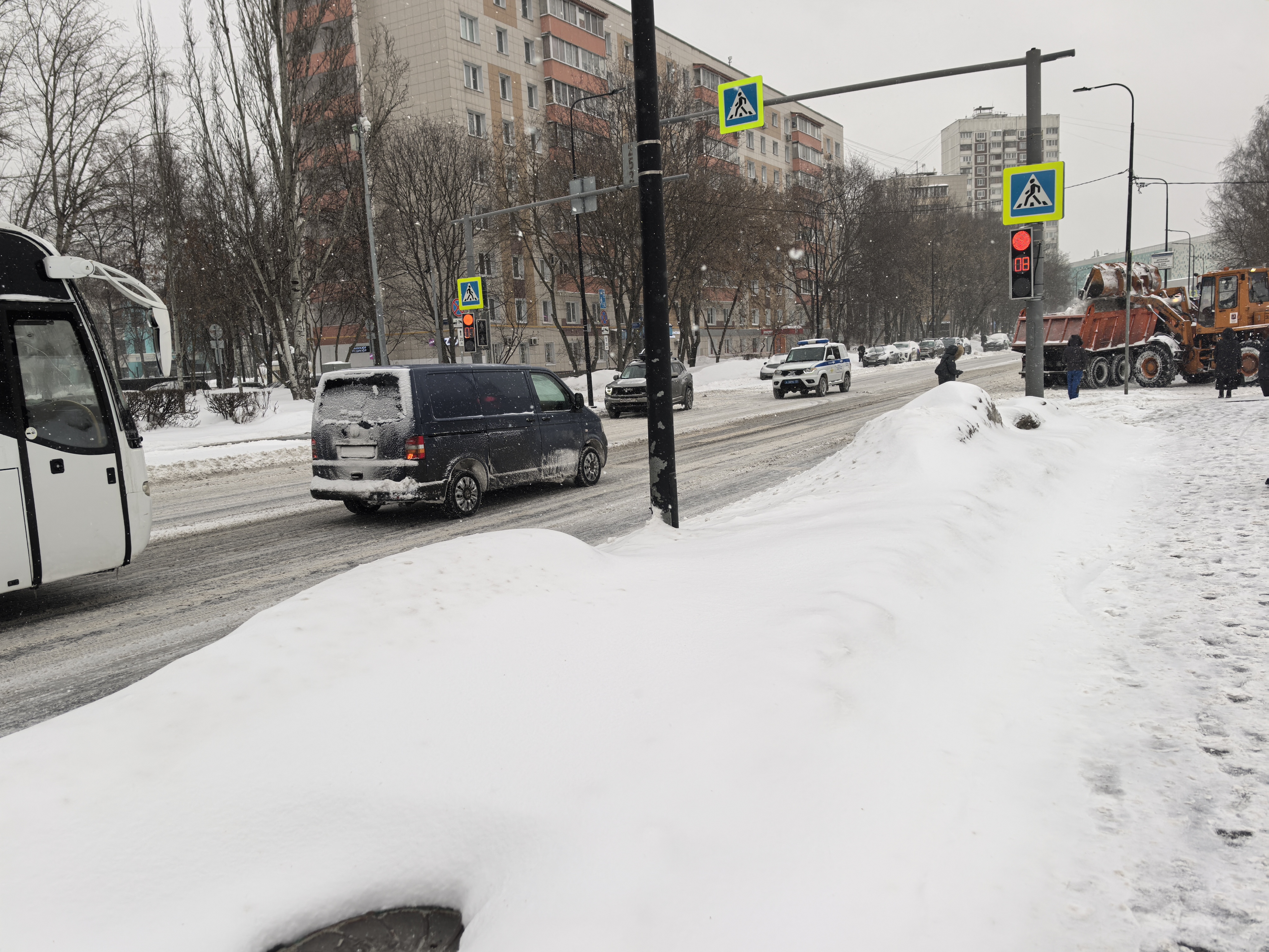 Городская улица с пешеходным переходом, регулируемым светофором. На дороге снег и слякоть, видны движущиеся транспортные средства.