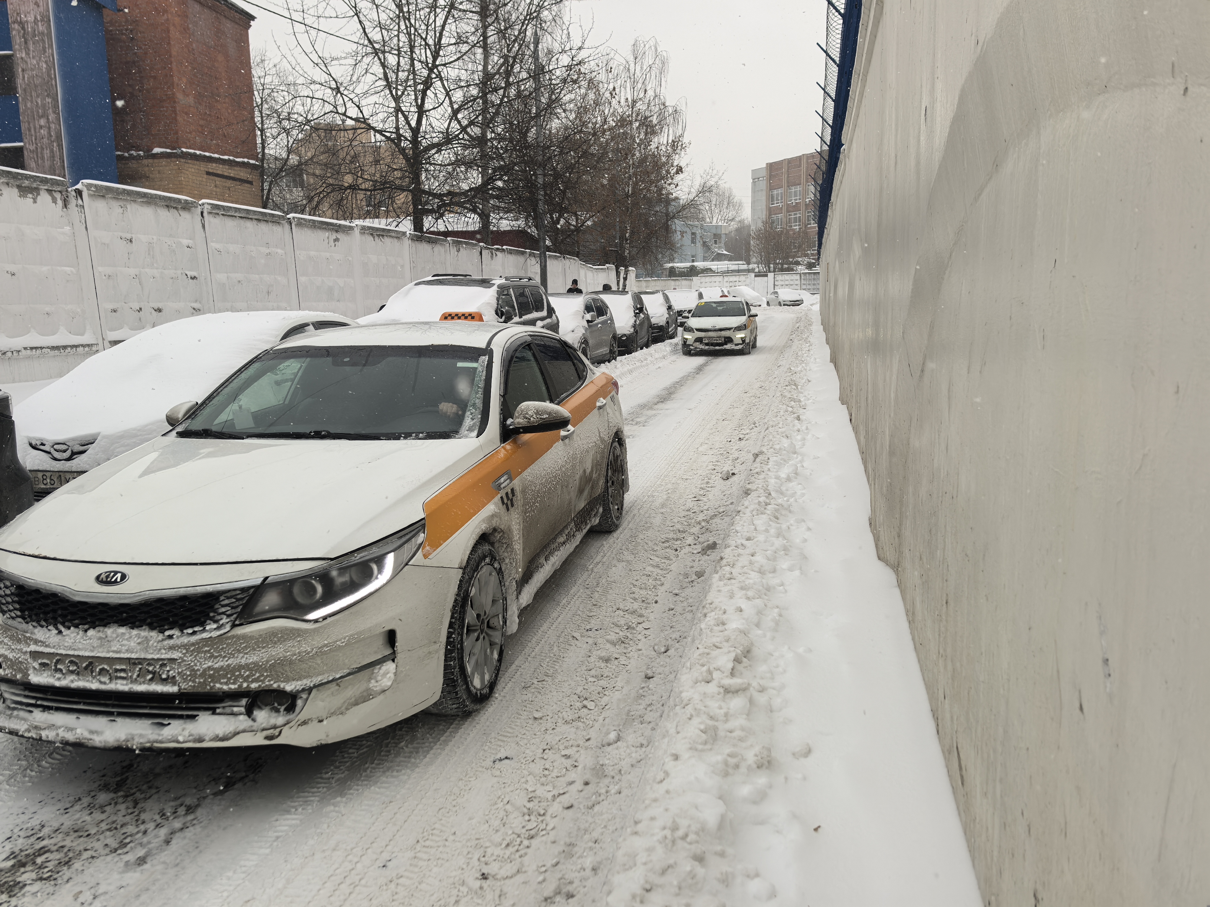 Узкая проезжая часть в зимних условиях, покрытая снегом и наледью. Тротуар отсутствует, пешеходы вынуждены идти по проезжей части между припаркованными машинами и стеной. Переходов нет.