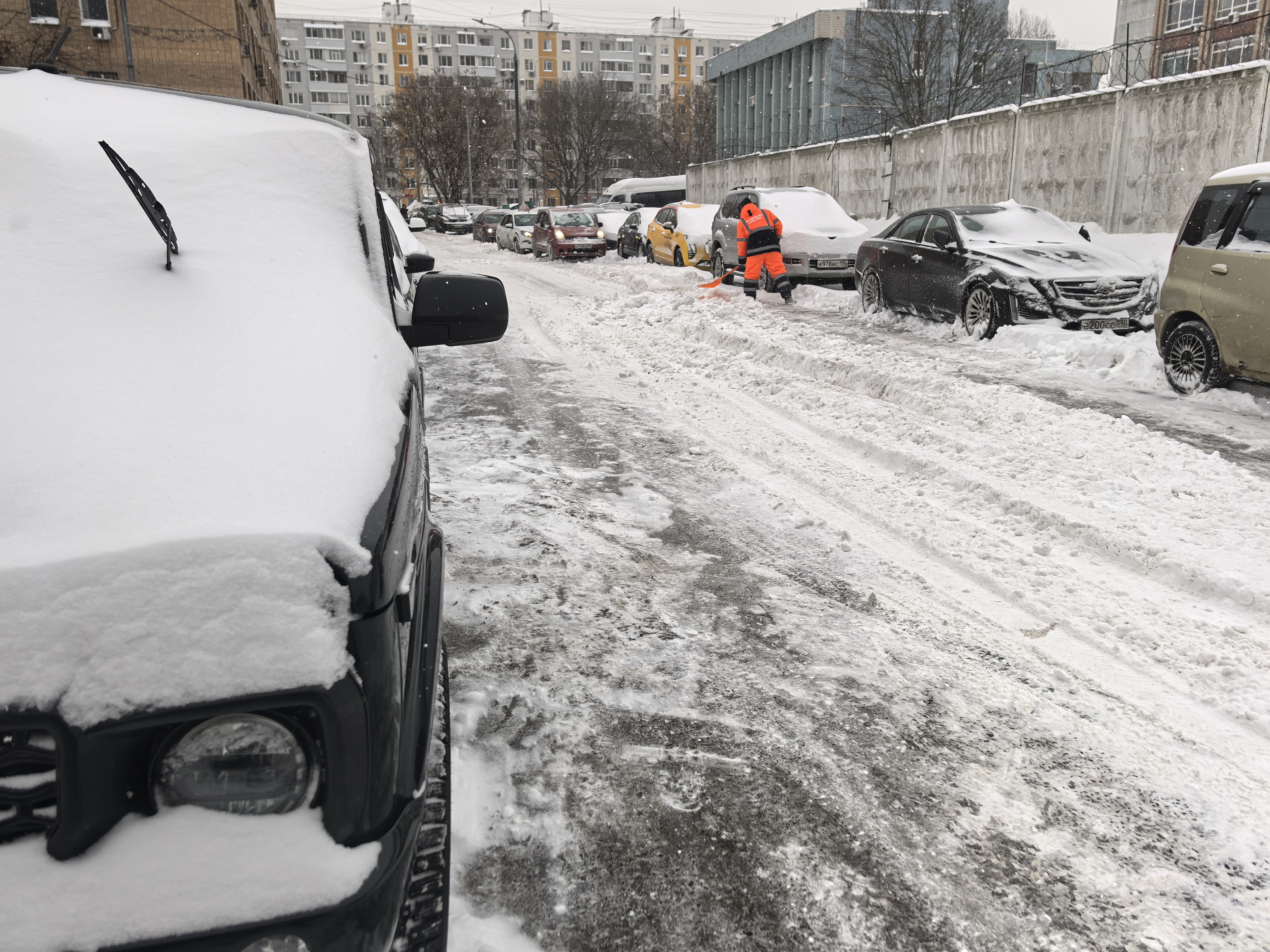 Городская улица в зимний период, покрытая снегом и слякотью. Пешеходный переход отсутствует, тротуары не видны или полностью засыпаны снегом, что вынуждает пешеходов идти по проезжей части. Парковка автомобилей вдоль дороги блокирует обзор и сужает проезж