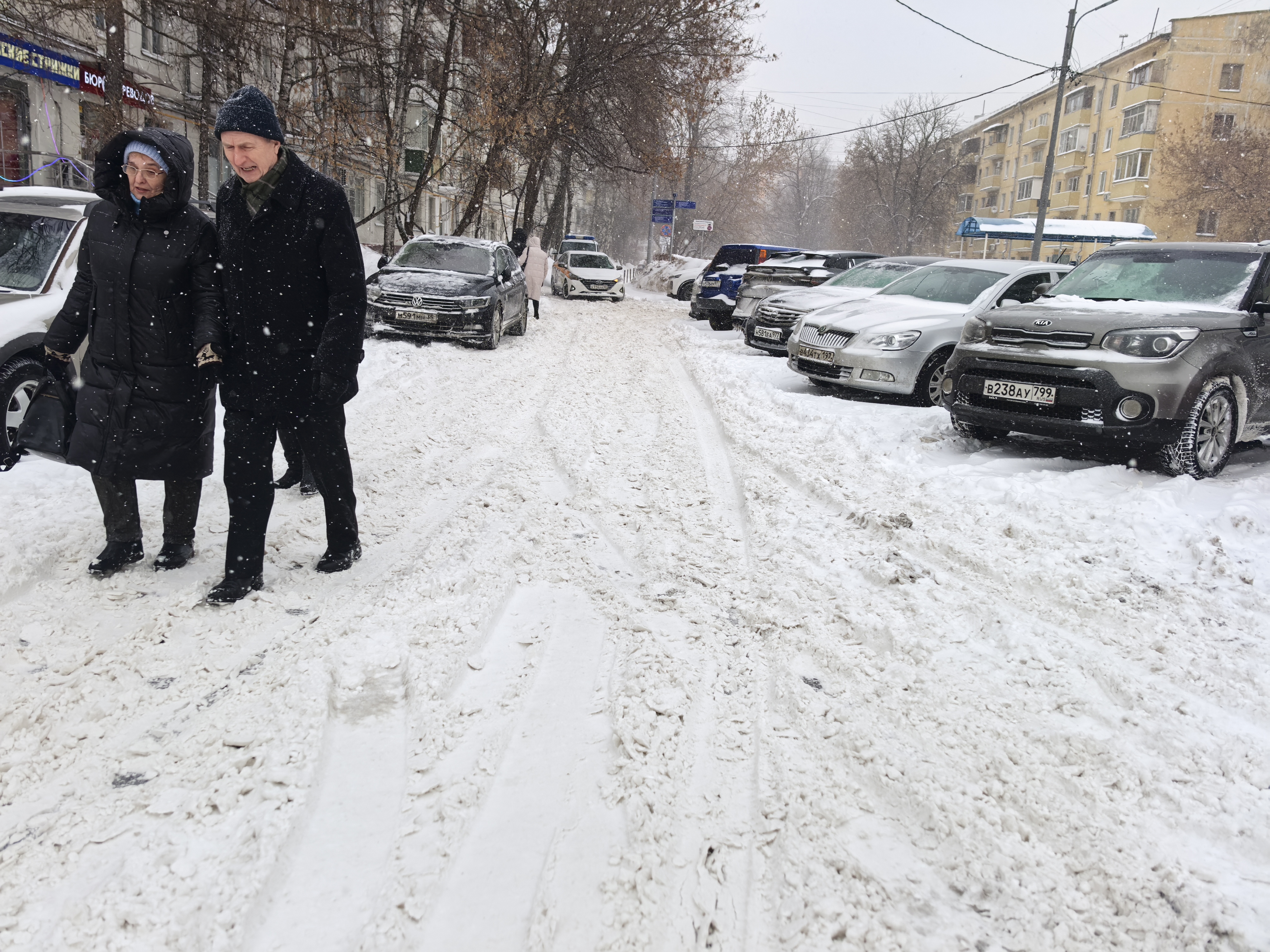 Участок городской улицы в зимний период, покрытый снегом. Пешеходный переход (зебра) отсутствует. Парковка автомобилей вдоль дороги, тротуары не просматриваются или полностью засыпаны снегом.