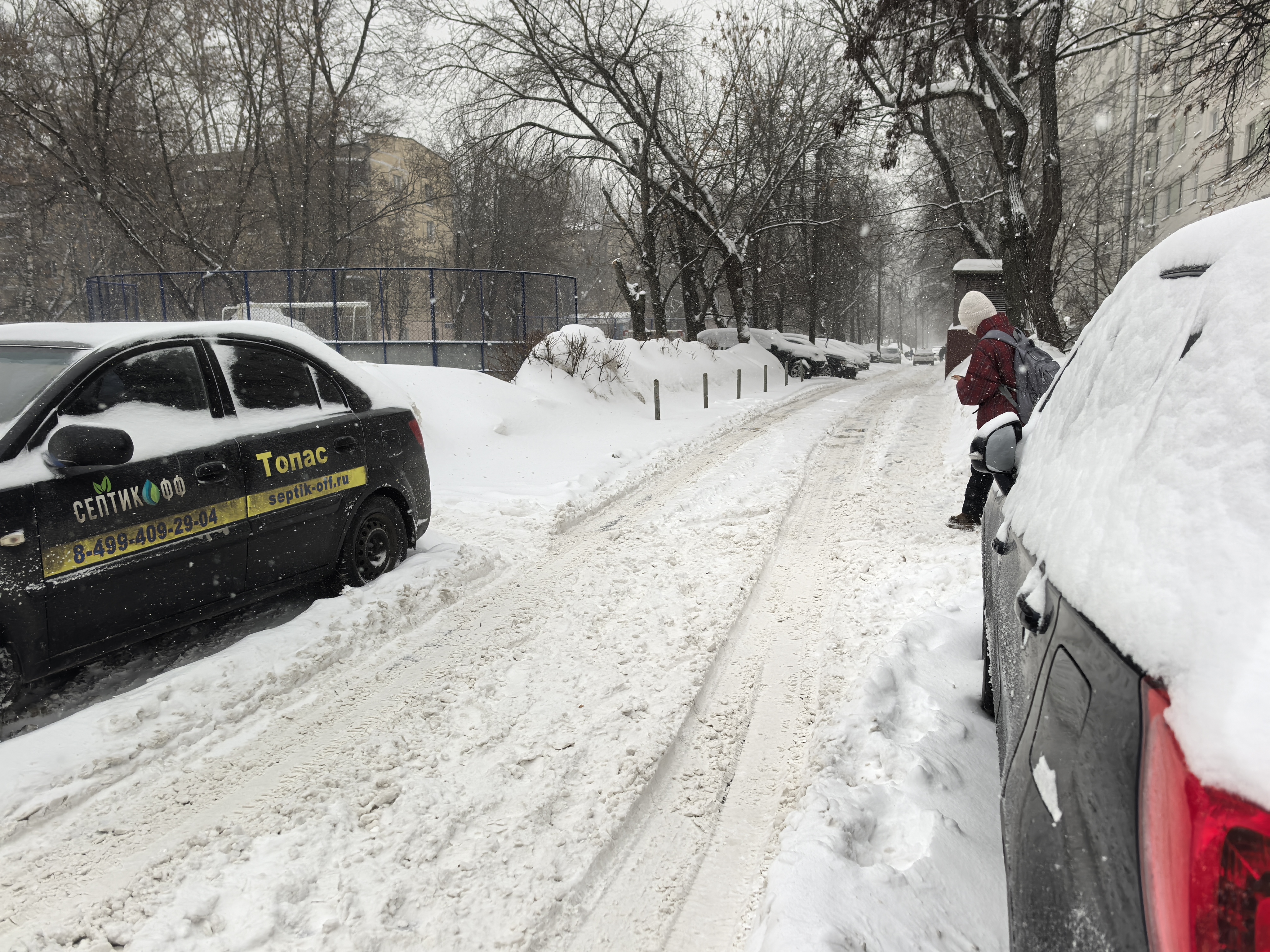 Участок дороги в зимний период, покрытый снегом и наледью. Тротуары полностью засыпаны снегом, пешеход вынужден идти по проезжей части. Видна припаркованная техника, ограничивающая обзор и проход.