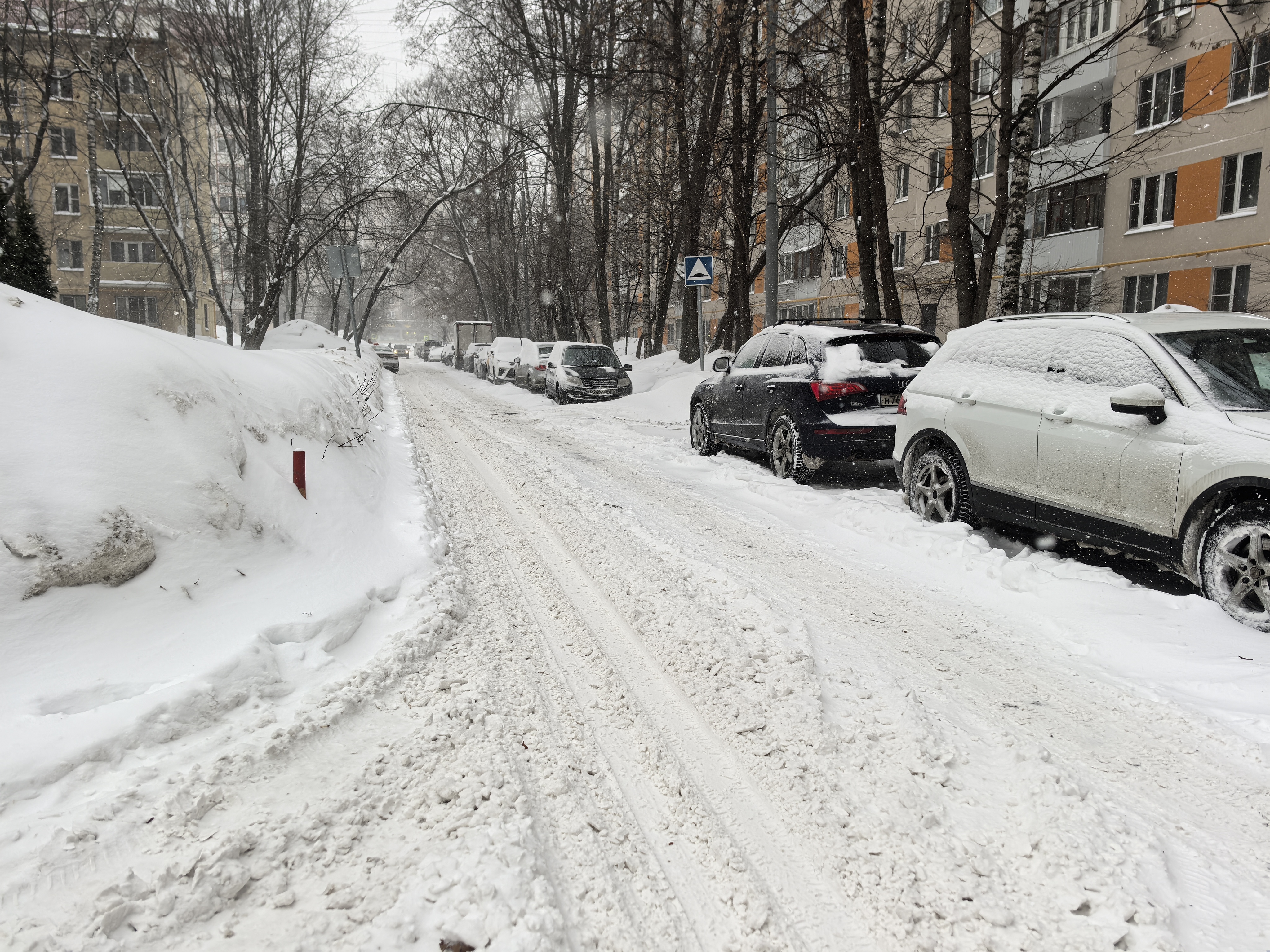Участок проезжей части в жилом районе, покрытый снегом. Пешеходный переход отсутствует, но виден знак, предупреждающий о его приближении впереди. Тротуары полностью засыпаны снегом.