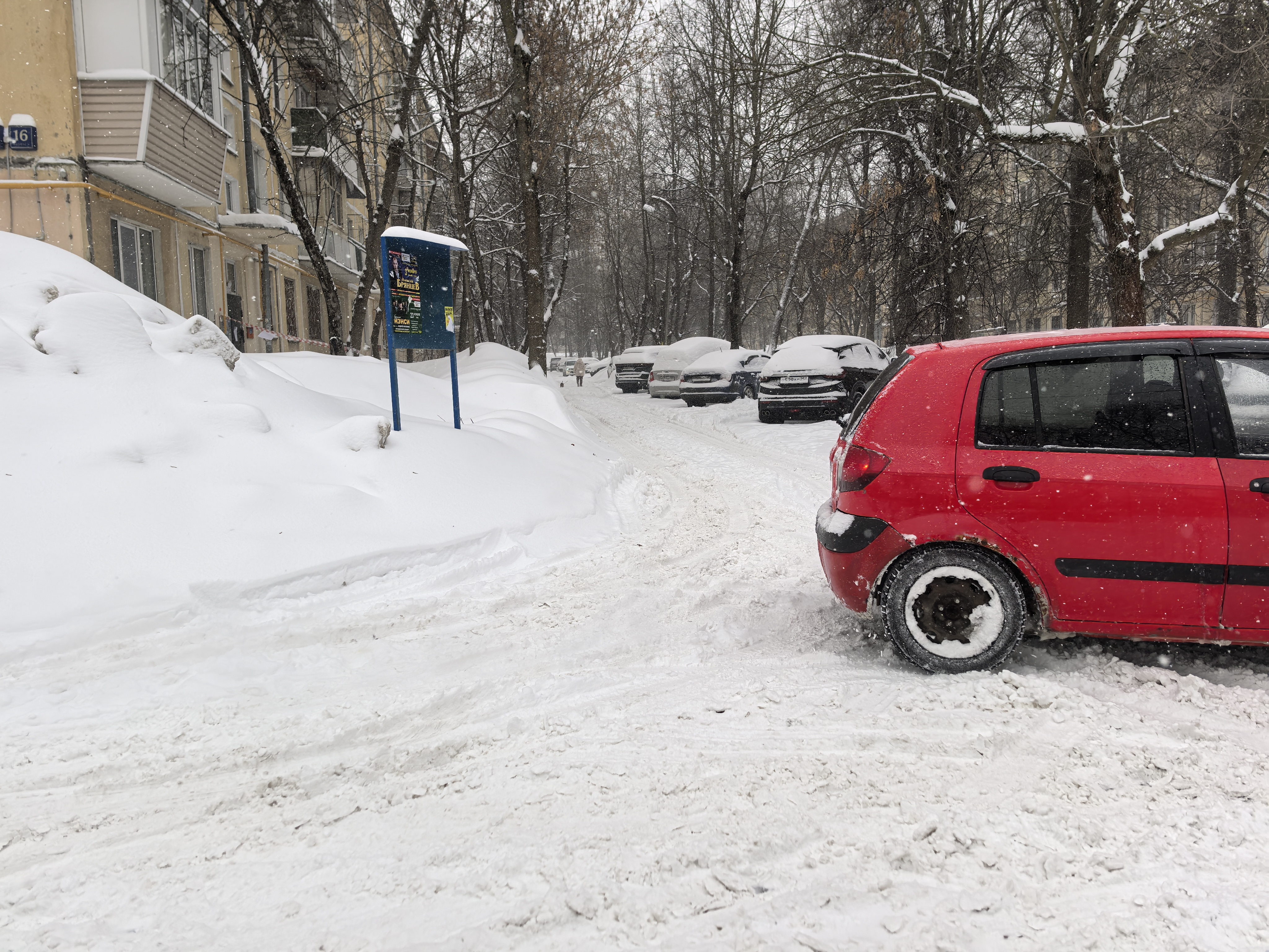 Дворовая территория или прилегающая к дому дорога, полностью покрытая снегом. Видны припаркованные автомобили, тротуары не расчищены.