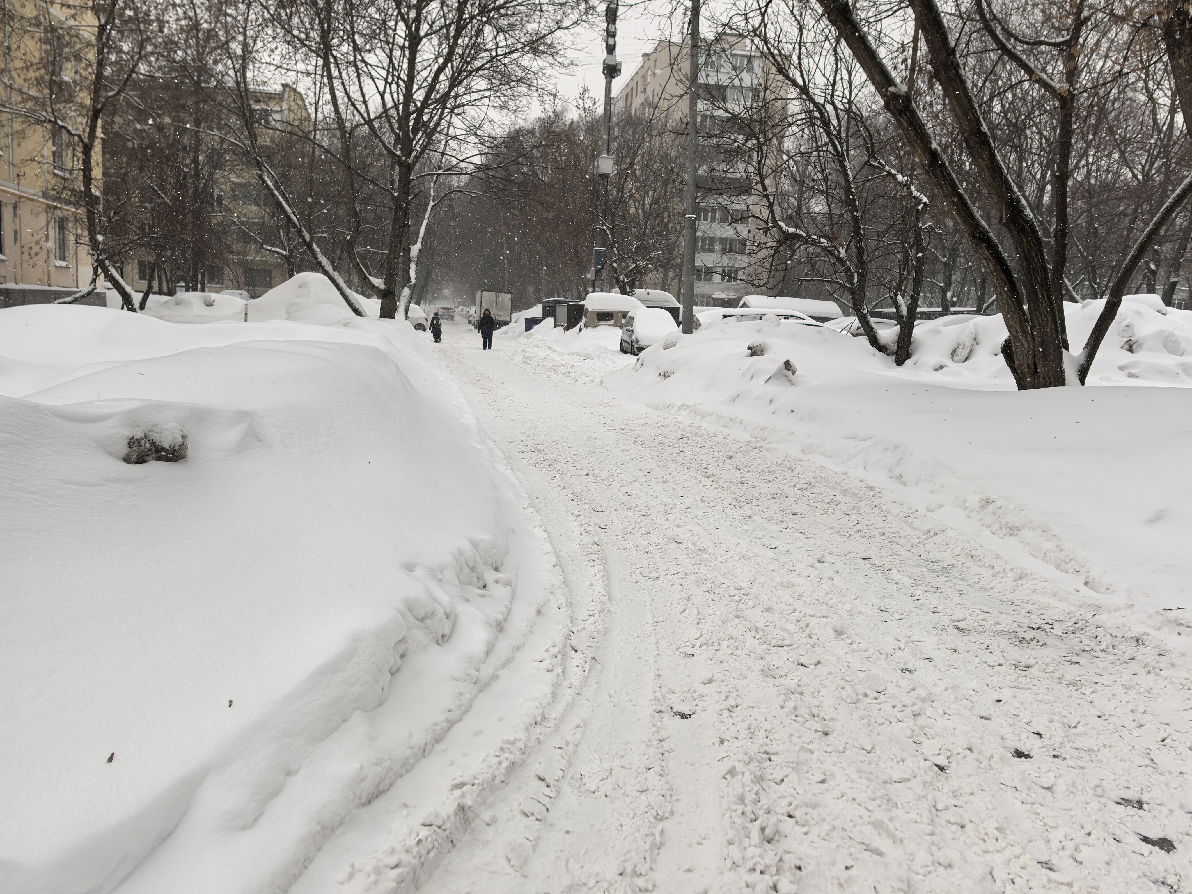 Дворовая территория, покрытая снегом. Видны припаркованные автомобили, засыпанные снегом, и следы от шин. Пешеходы идут по проезжей части, так как тротуары полностью засыпаны снегом.