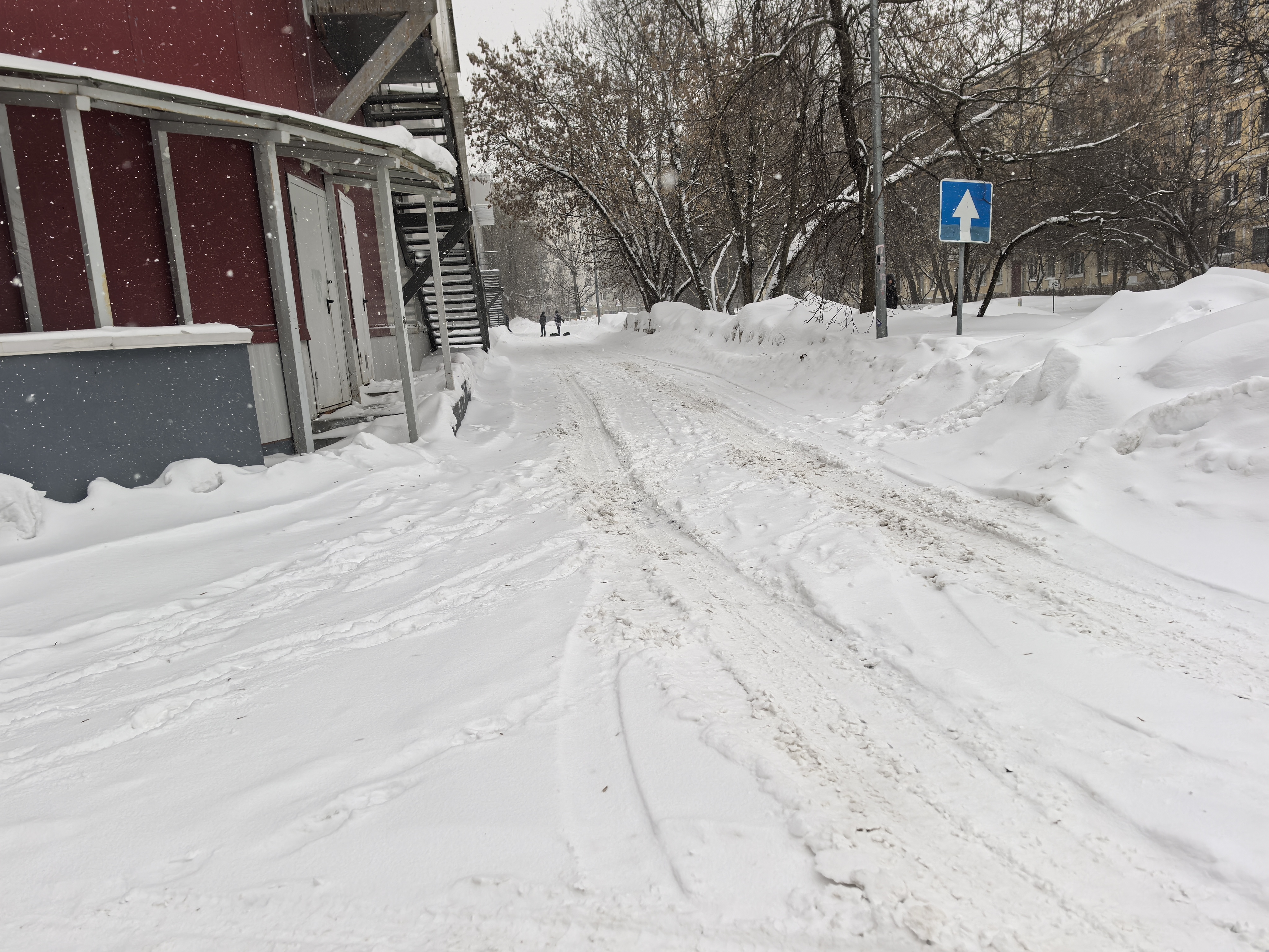 Заснеженная дворовая территория или подъездная дорога к зданию. Дорога полностью покрыта снегом, бордюры и тротуары не просматриваются. Видны следы от шин, что указывает на движение транспорта.
