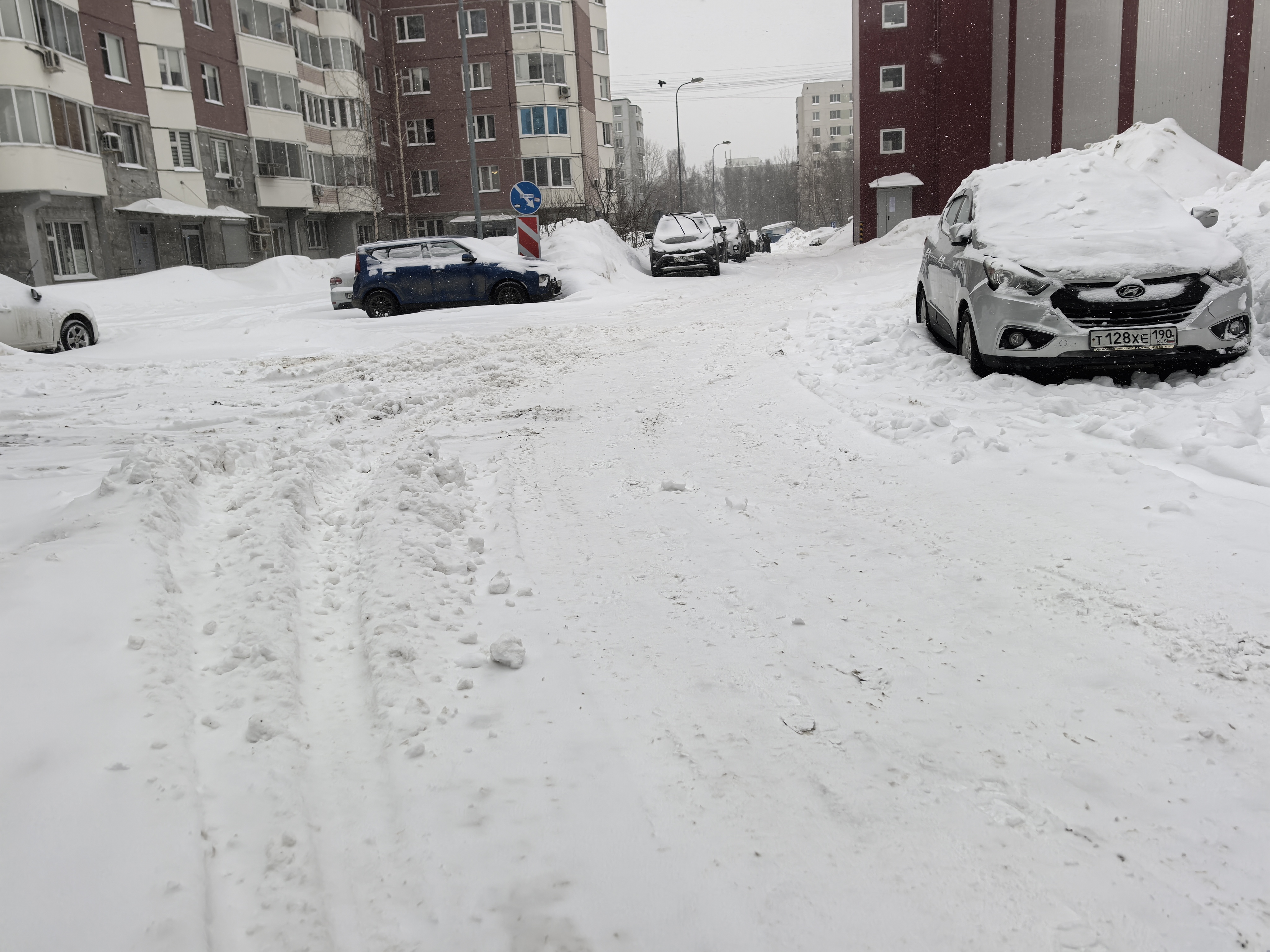 Дворовая территория между жилыми домами, покрытая снегом. Видны припаркованные автомобили и отсутствие пешеходных дорожек.