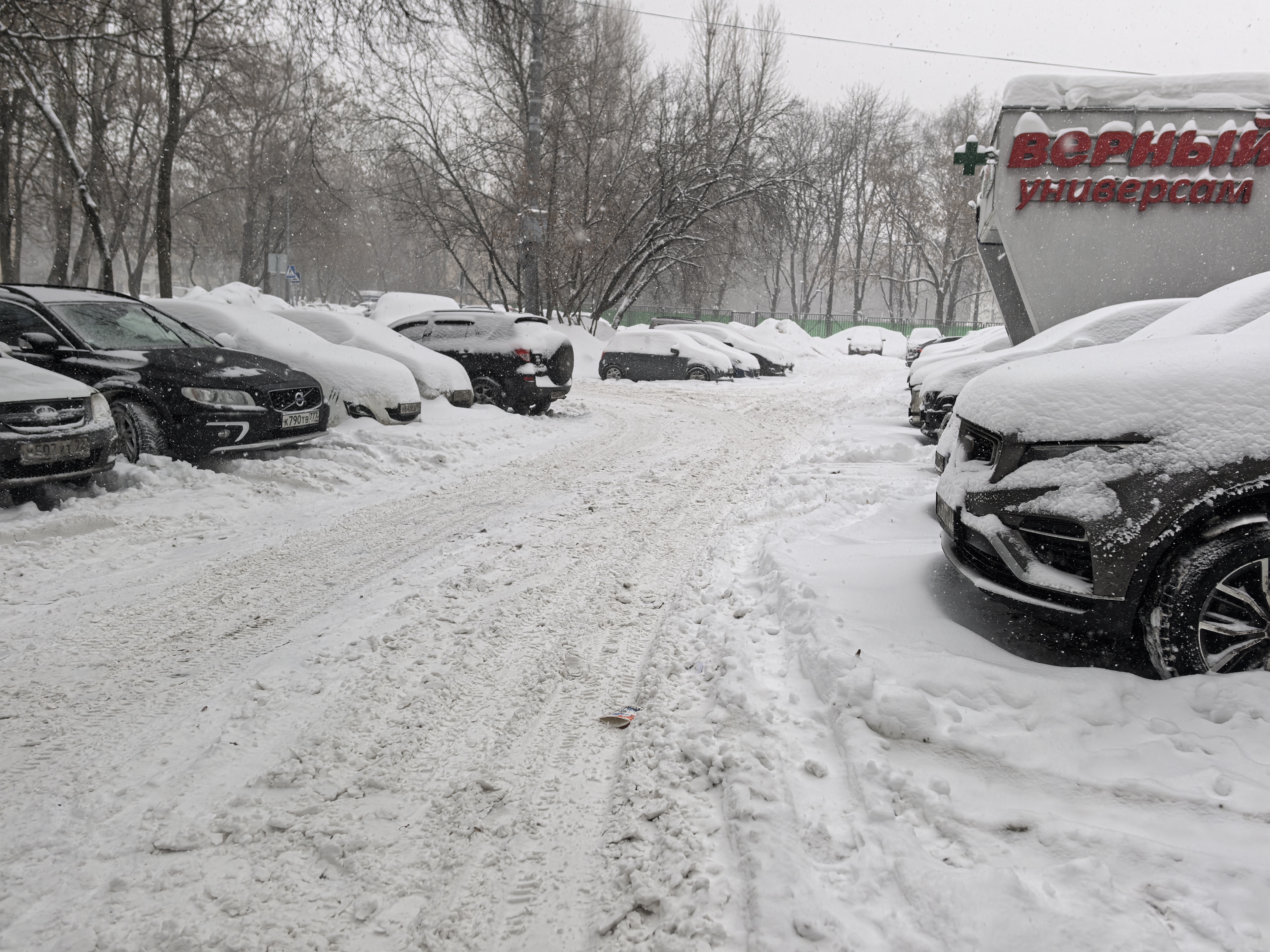 Дворовая территория или парковка у магазина, покрытая снегом. Видны припаркованные автомобили, дорожная разметка отсутствует, пешеходных зон не видно.