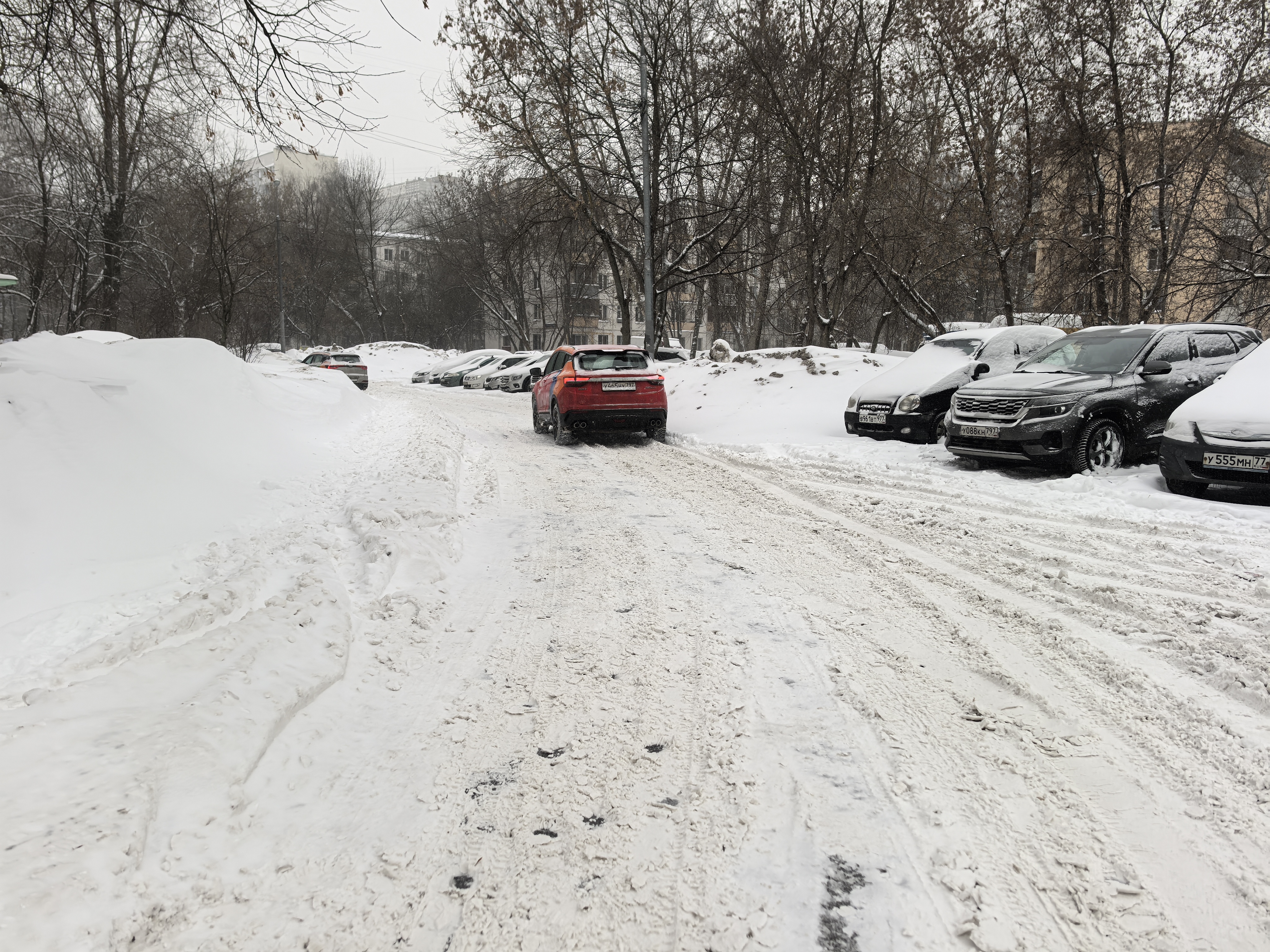Дворовая территория, покрытая снегом, с припаркованными автомобилями и проезжающим транспортом. Пешеходных зон и разметки не видно.