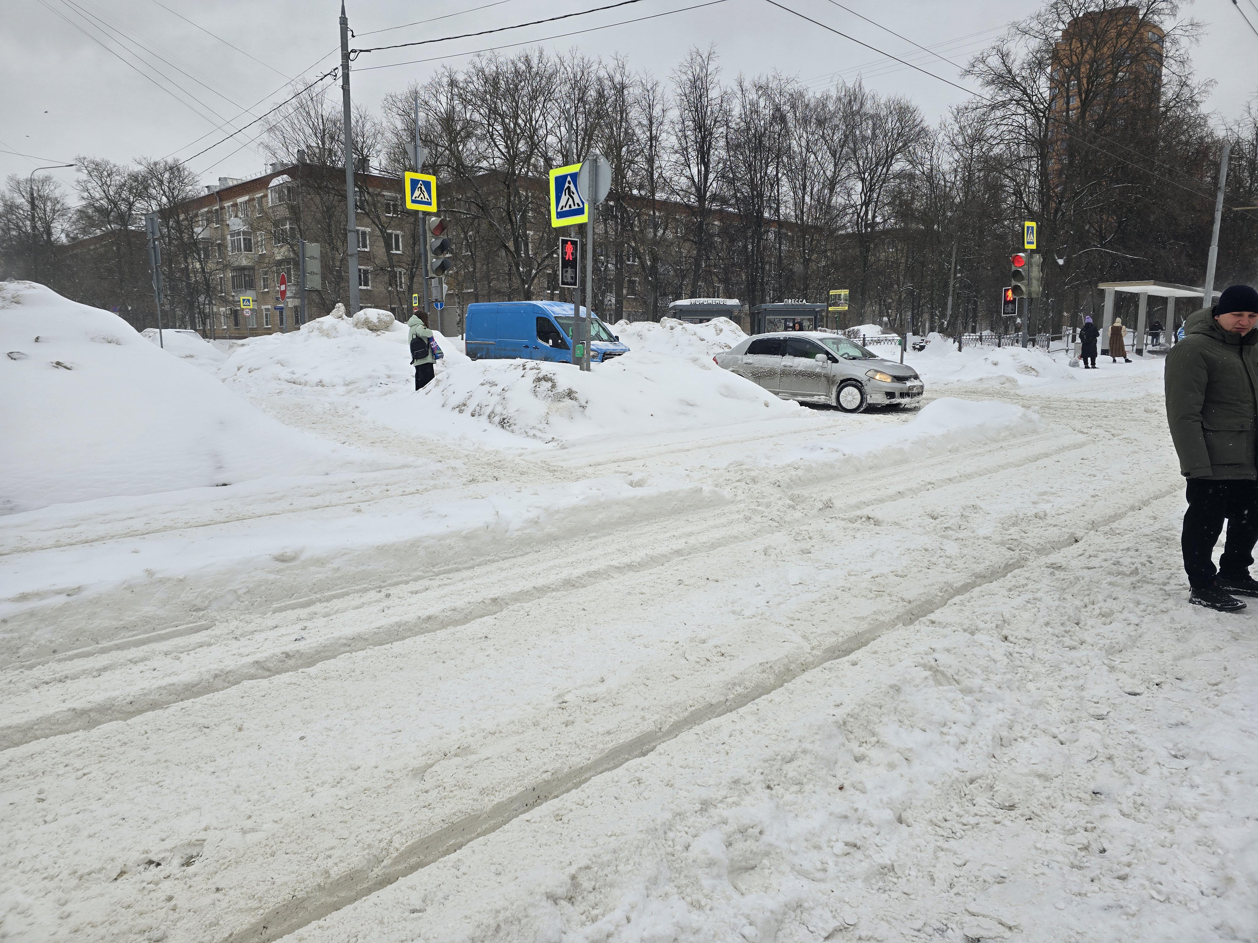 Проезжая часть с пешеходным переходом в зимних условиях. Видны светофоры, знаки перехода, но вся дорога и обочины покрыты снегом, разметка скрыта.