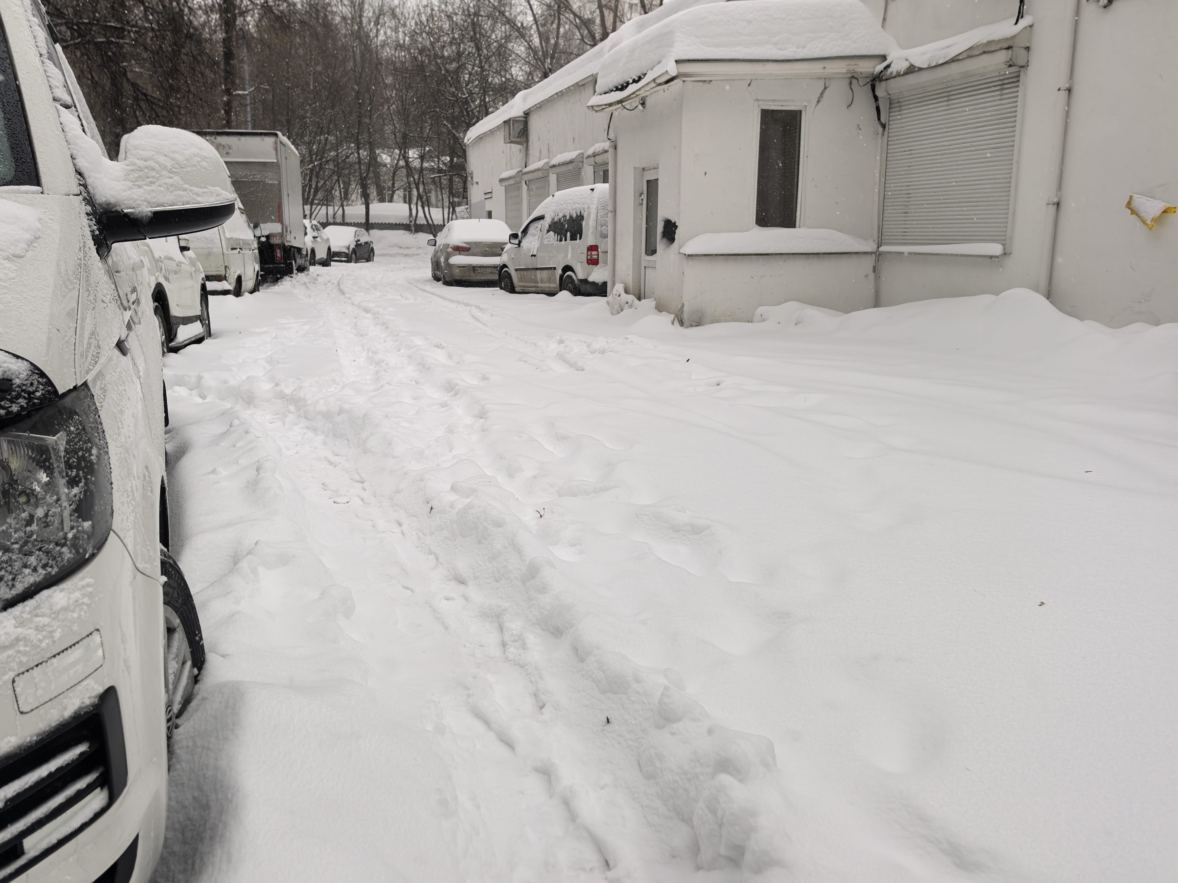 Дворовая территория, полностью покрытая снегом, с припаркованными автомобилями и зданиями по бокам. Проезжая часть не расчищена, видны только следы от шин.