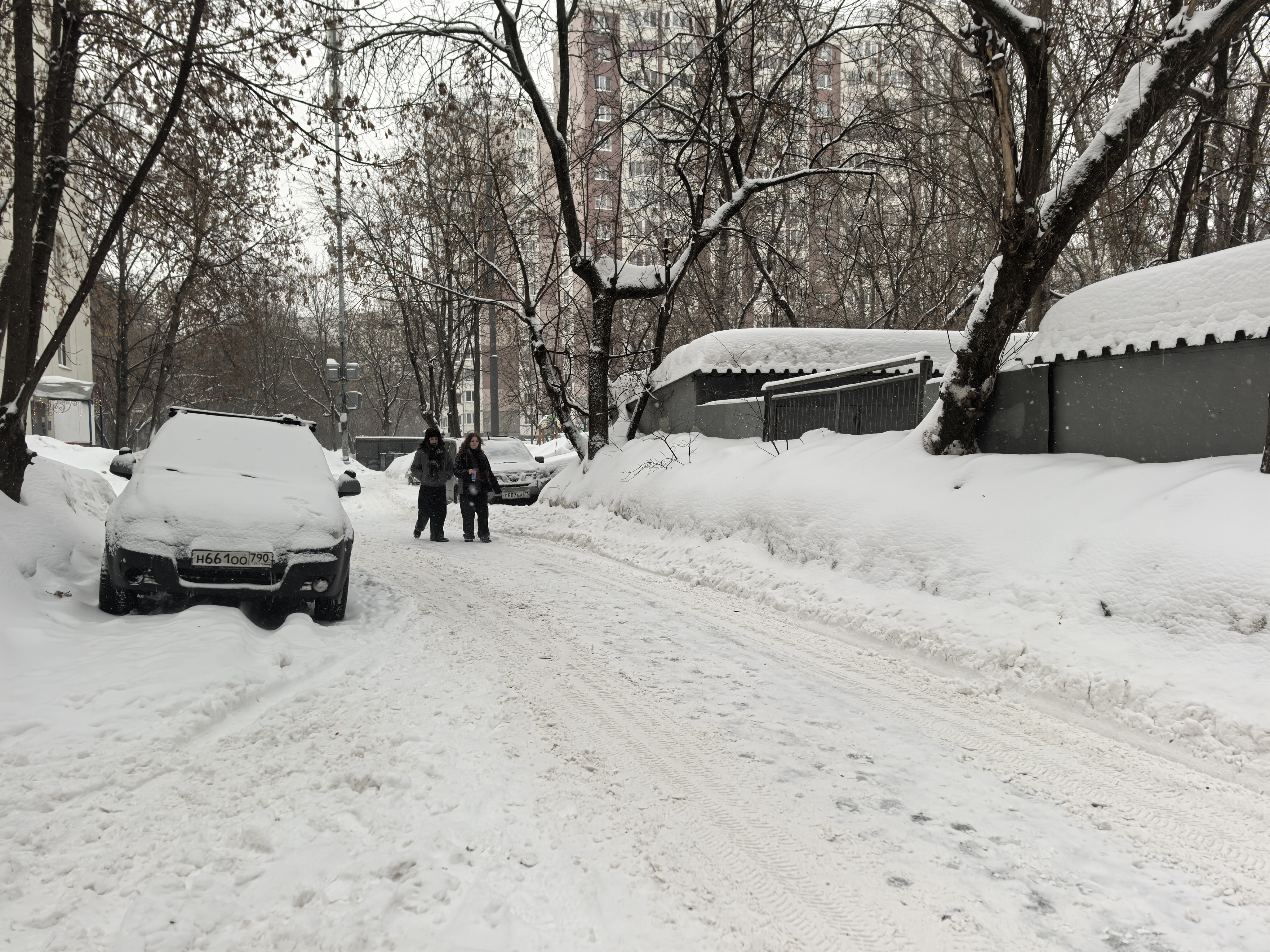 Дворовая территория с проезжей частью, покрытой снегом. Видны припаркованные автомобили и пешеходы, идущие по дороге. Вокруг деревья и жилые дома.
