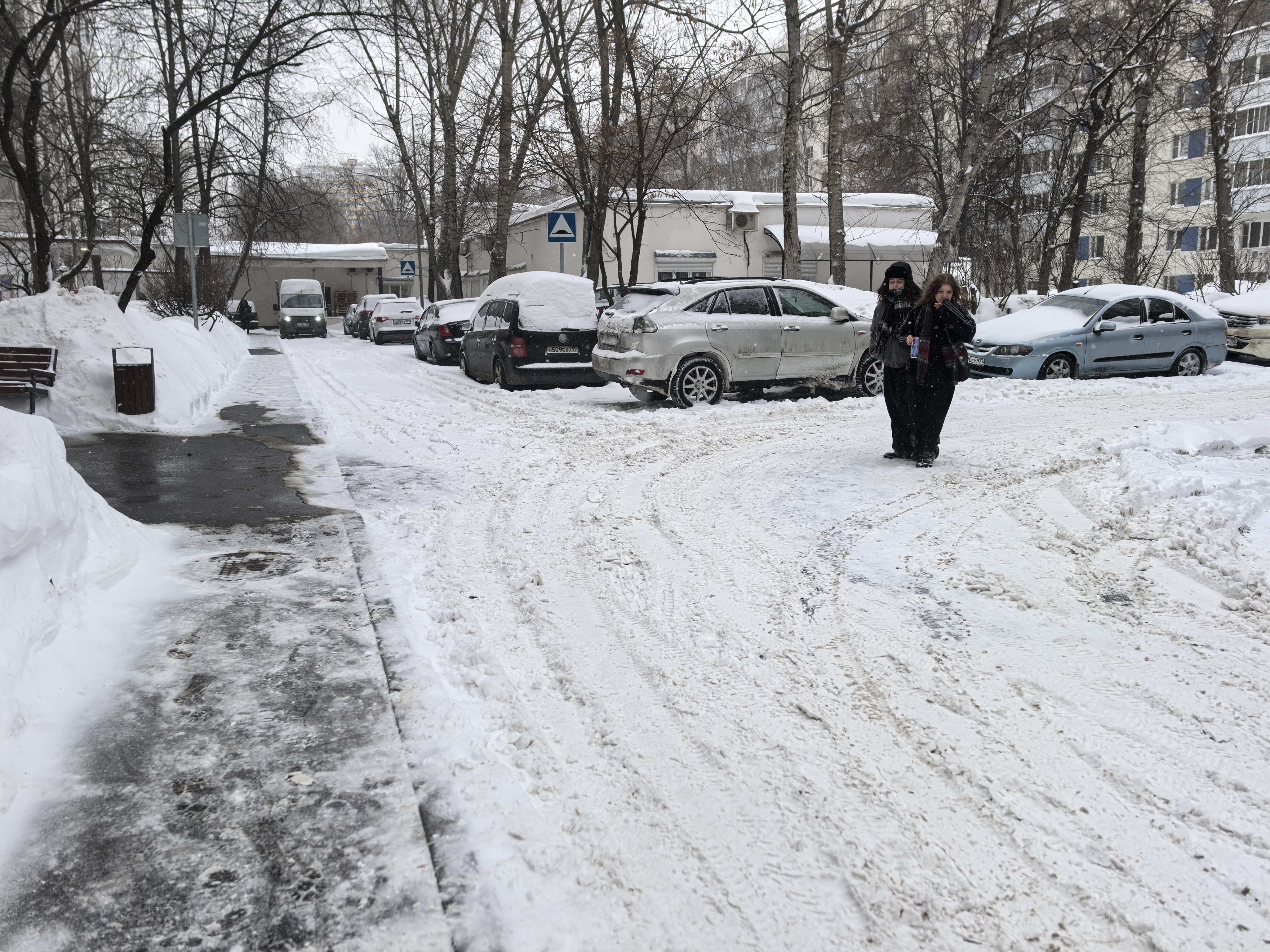 Дворовая территория с припаркованными автомобилями, покрытая снегом. Видны дети, идущие по проезжей части, и отсутствие четкого разделения на тротуар и дорогу.