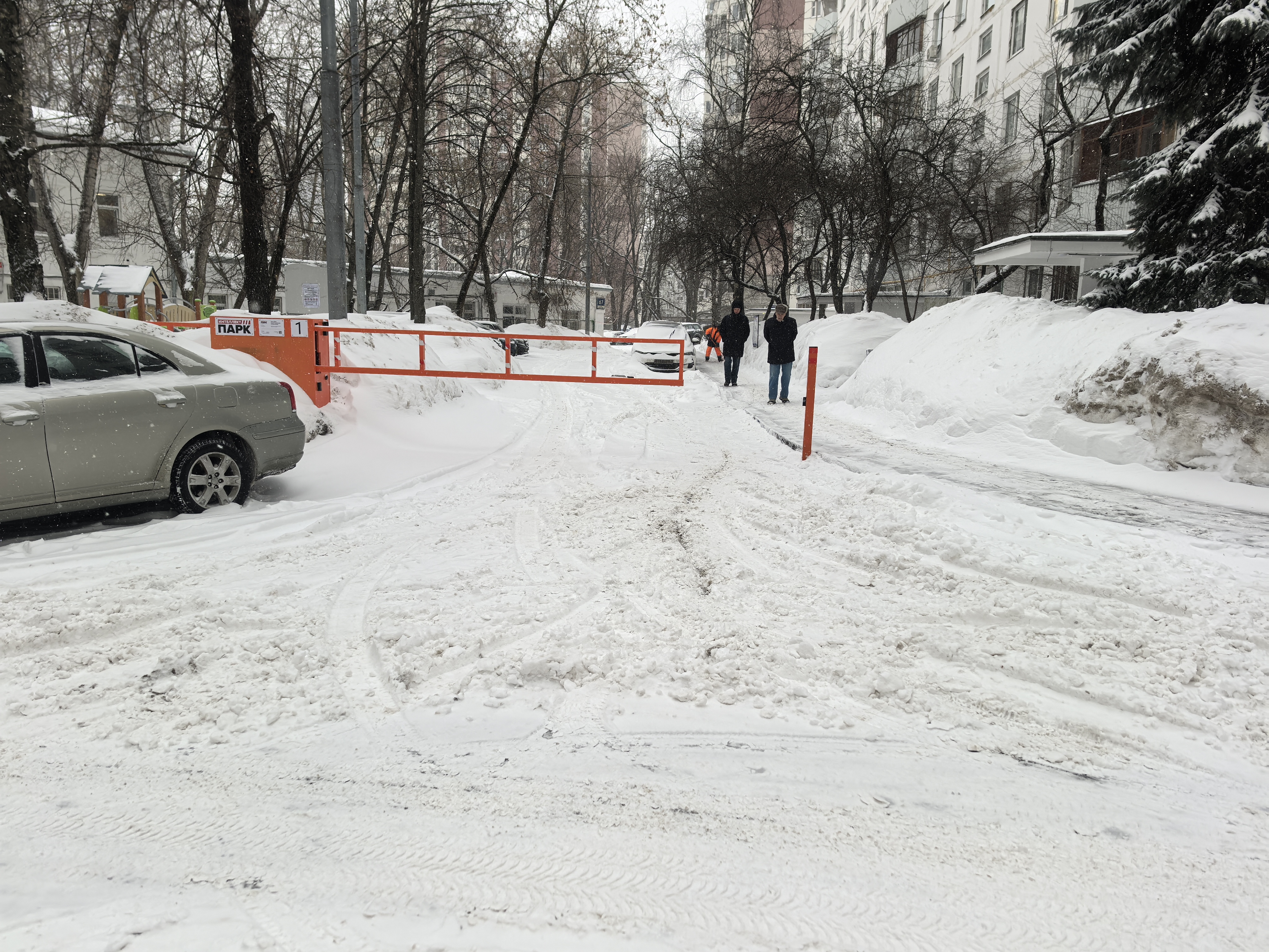 Въезд на закрытую парковку во дворе, полностью покрытый снегом. Видны припаркованные автомобили и пешеходы, идущие по снежной дороге.