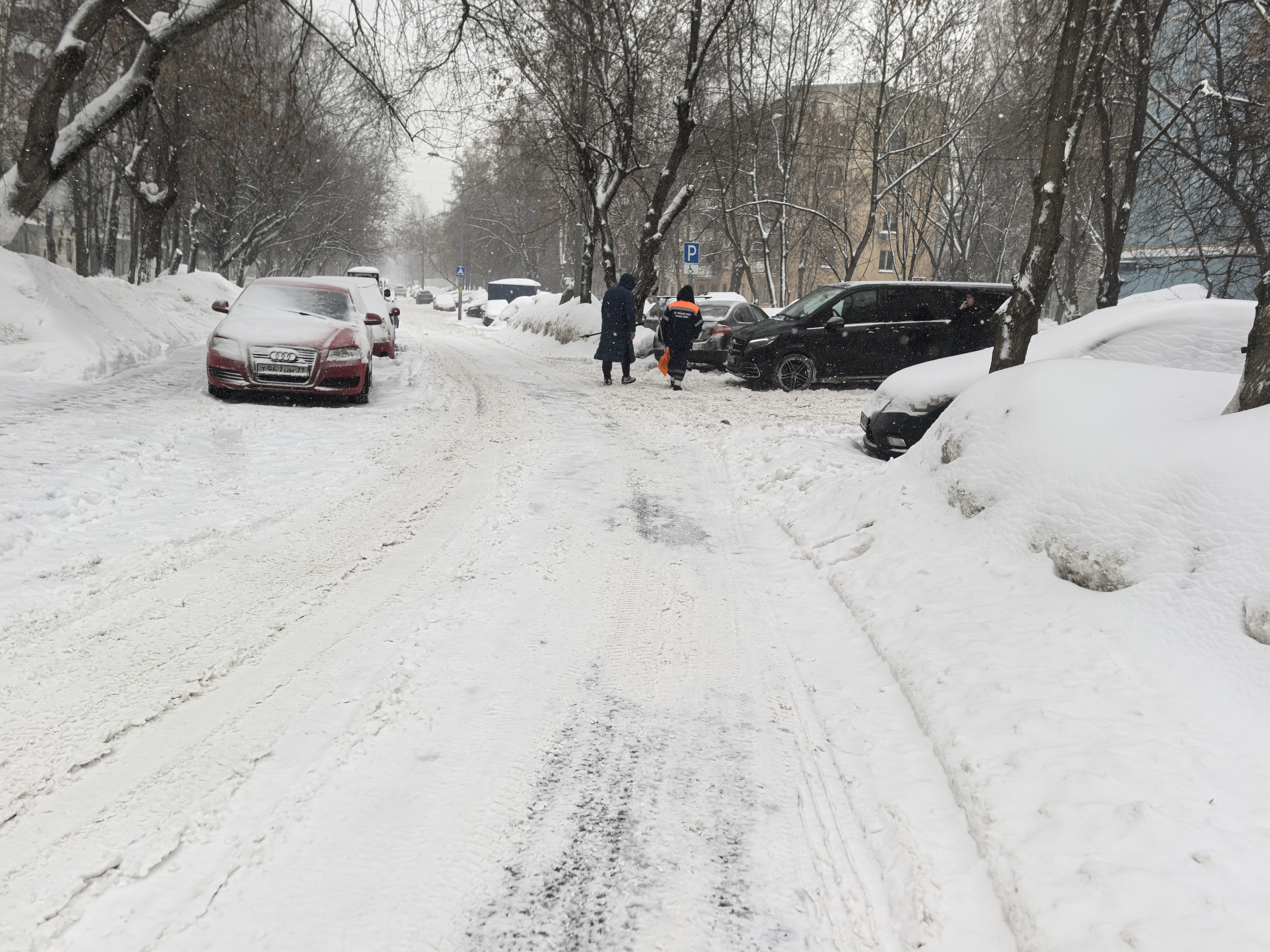 Участок городской улицы в зимний период. Дорога полностью покрыта снегом, разметка не видна. Пешеходный переход отсутствует. Тротуары полностью засыпаны снегом, пешеходы вынуждены идти по проезжей части. Парковка автомобилей вдоль дороги блокирует обзор и