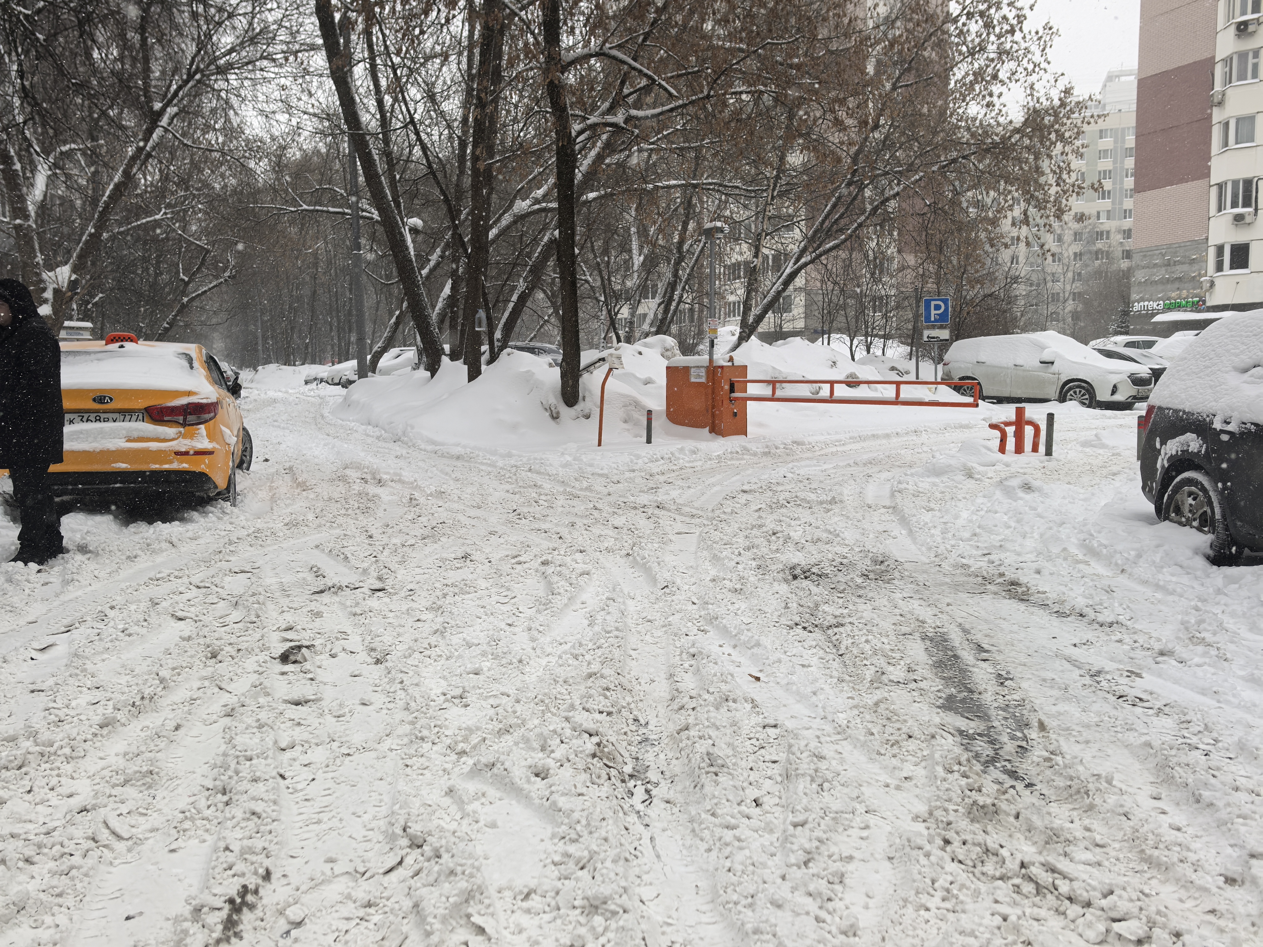 Дворовая территория с въездом на парковку, покрытая снегом. Видны припаркованные автомобили и шлагбаум.