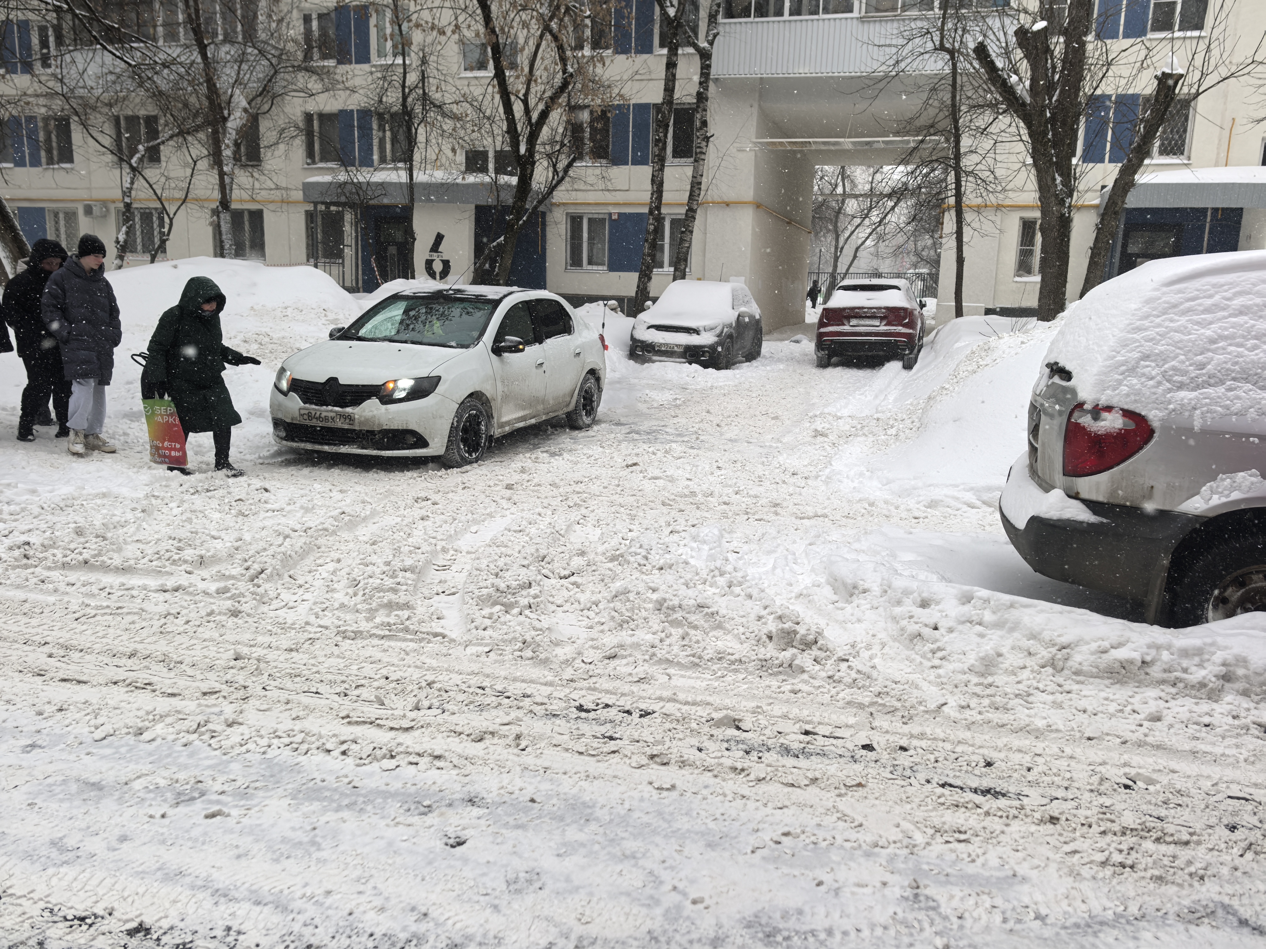 Дворовая территория жилого дома, покрытая снегом. Видны припаркованные автомобили и дети, идущие по проезжей части.