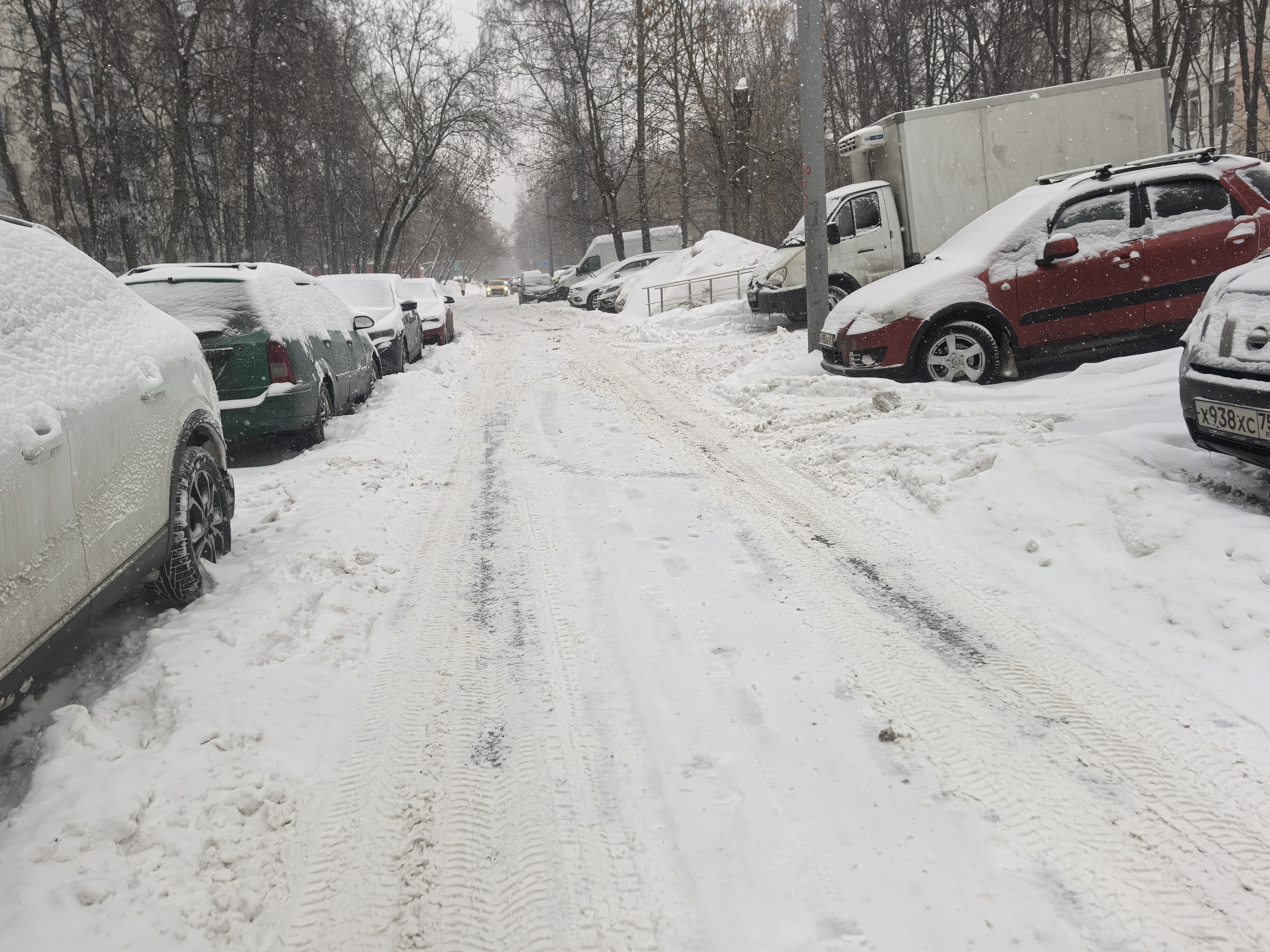 Участок проезжей части без пешеходного перехода, покрытый снегом, с припаркованными автомобилями по обеим сторонам. Тротуары не просматриваются или полностью скрыты под сугробами.