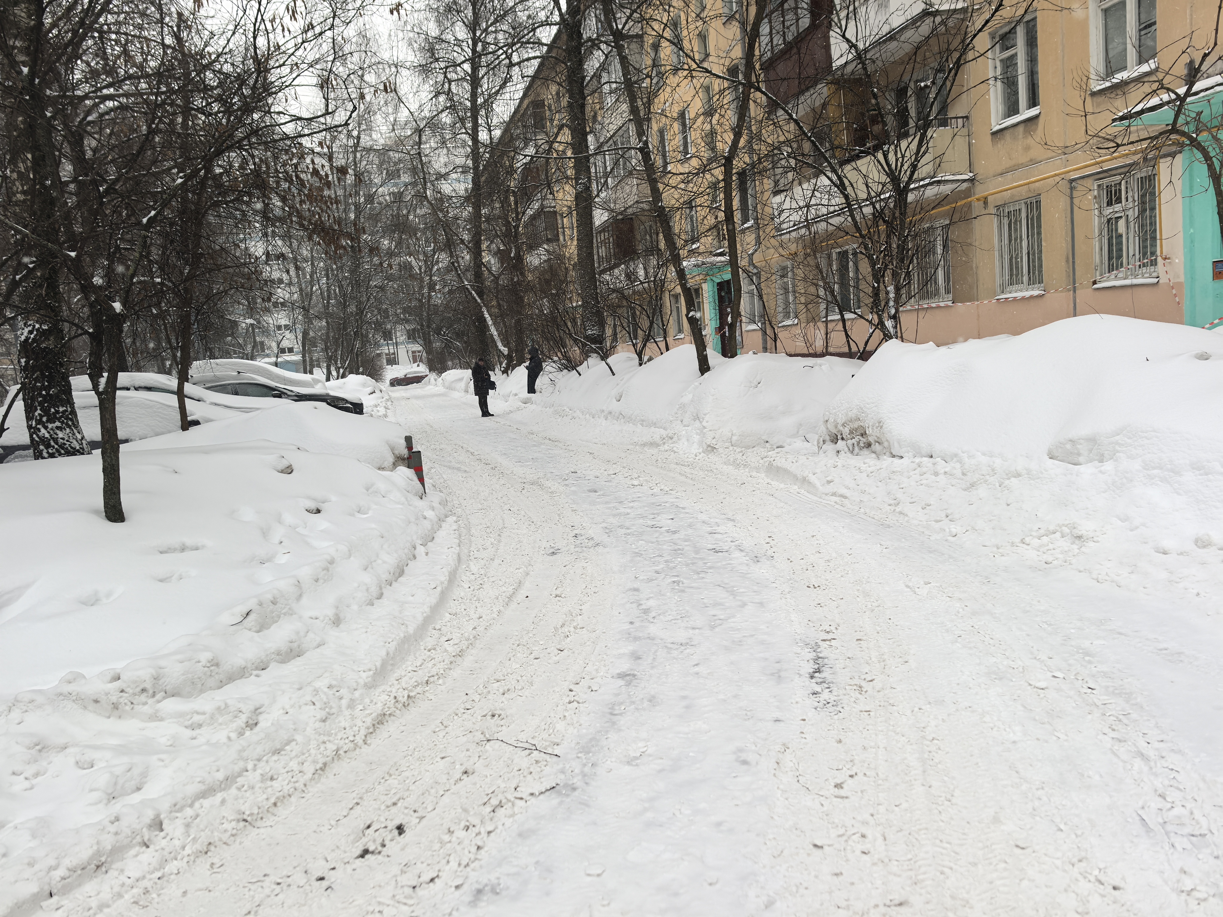 Дворовая территория с проезжей частью, покрытой снегом и следами от шин. Видны припаркованные автомобили и жилые дома.