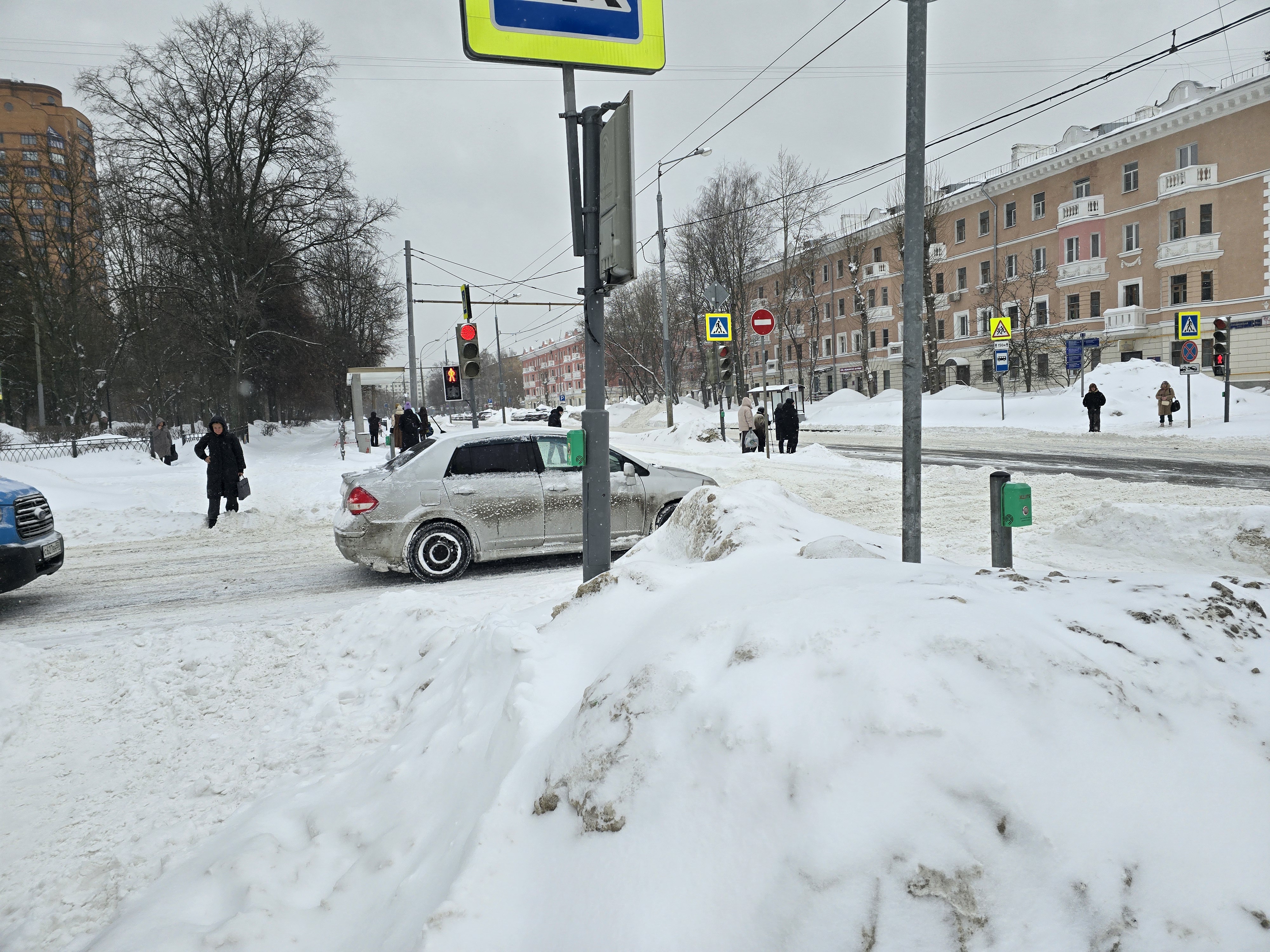 Городская улица с пешеходным переходом, регулируемым светофором, в зимних условиях. Видны здания, деревья и припаркованные автомобили.