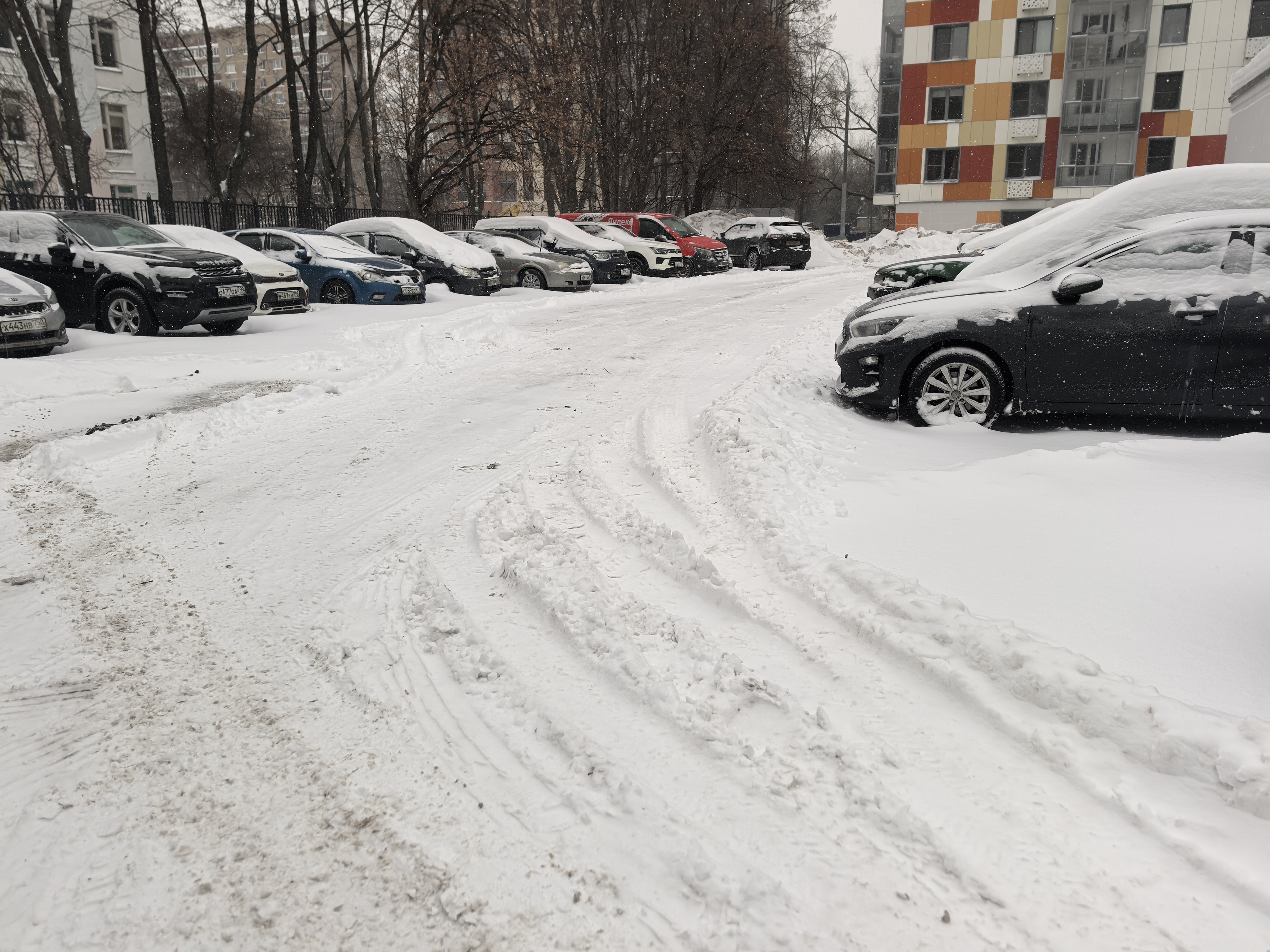 Дворовая территория с парковкой, покрытая снегом. Проезжая часть узкая, ограничена припаркованными автомобилями с обеих сторон. Пешеходных переходов и тротуаров не видно.