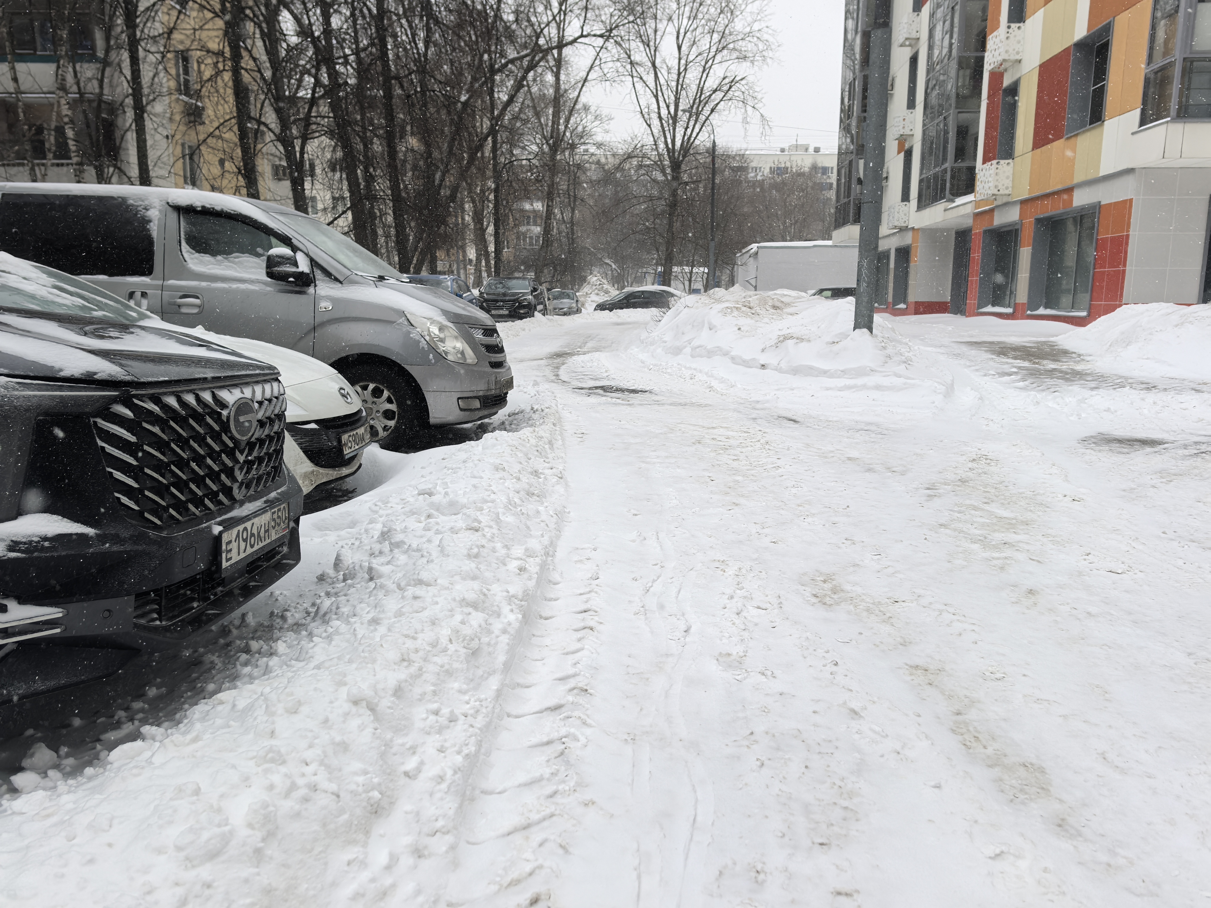 Дворовая территория с припаркованными автомобилями, покрытая снегом. Проезжая часть узкая, тротуаров нет, движение пешеходов происходит по снежной поверхности дороги.
