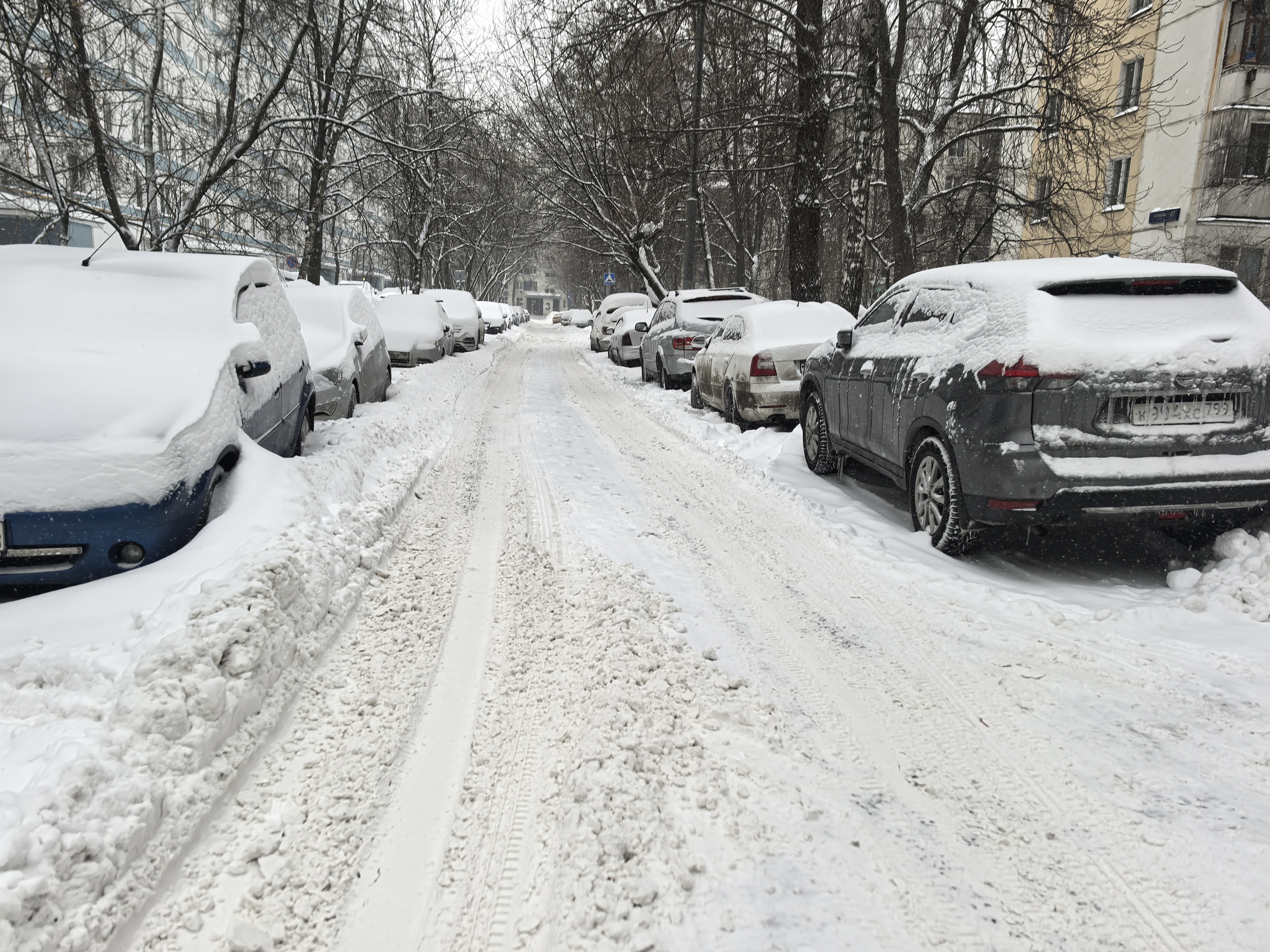 Дворовая территория, узкая проезжая часть, полностью покрытая снегом, с припаркованными автомобилями по обеим сторонам. Видны жилые дома и деревья.