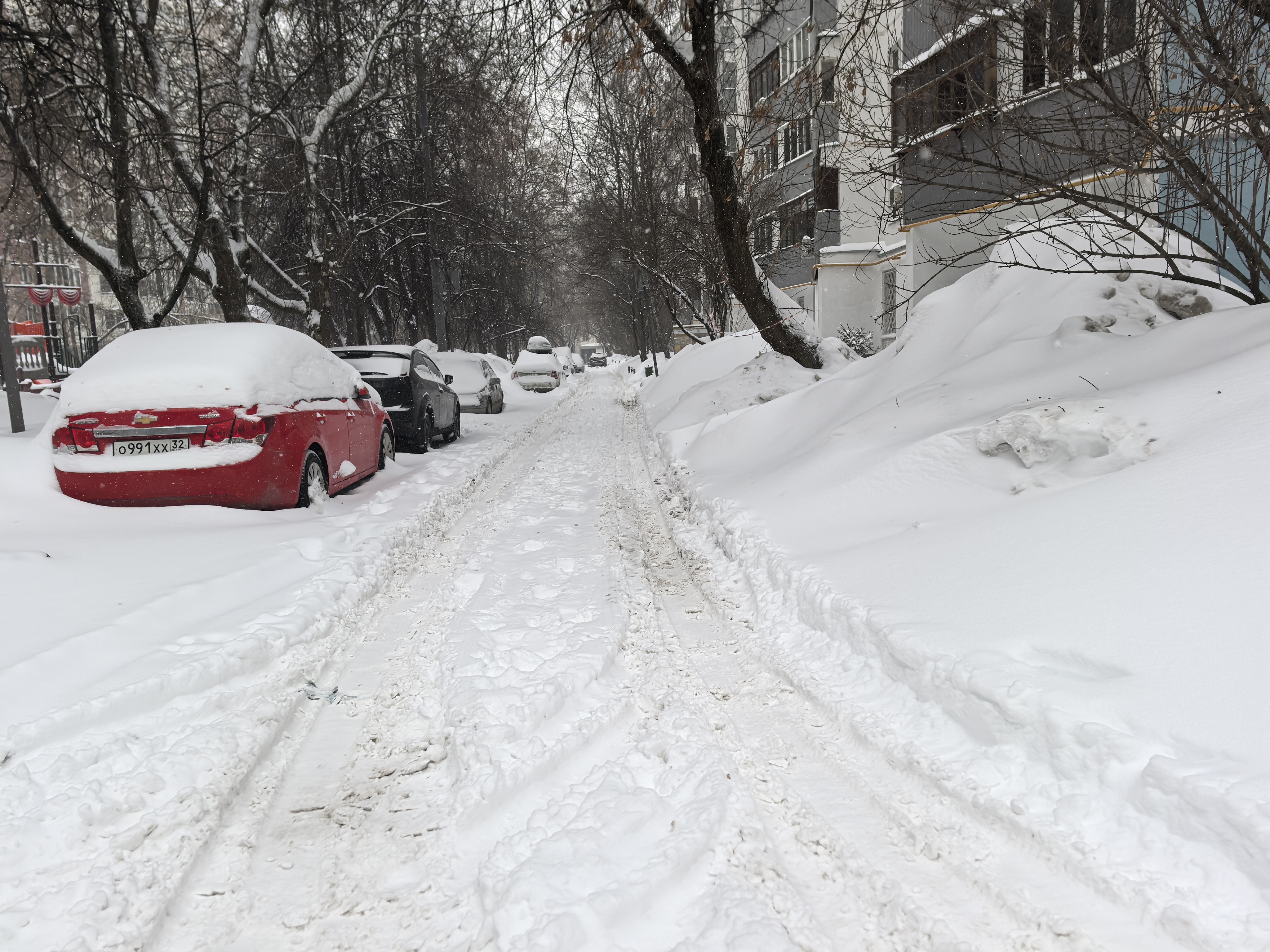 Дворовая территория, покрытая снегом. Видны припаркованные автомобили, жилой дом и детская площадка слева. Проезжая часть узкая, покрыта снегом и следами от шин.