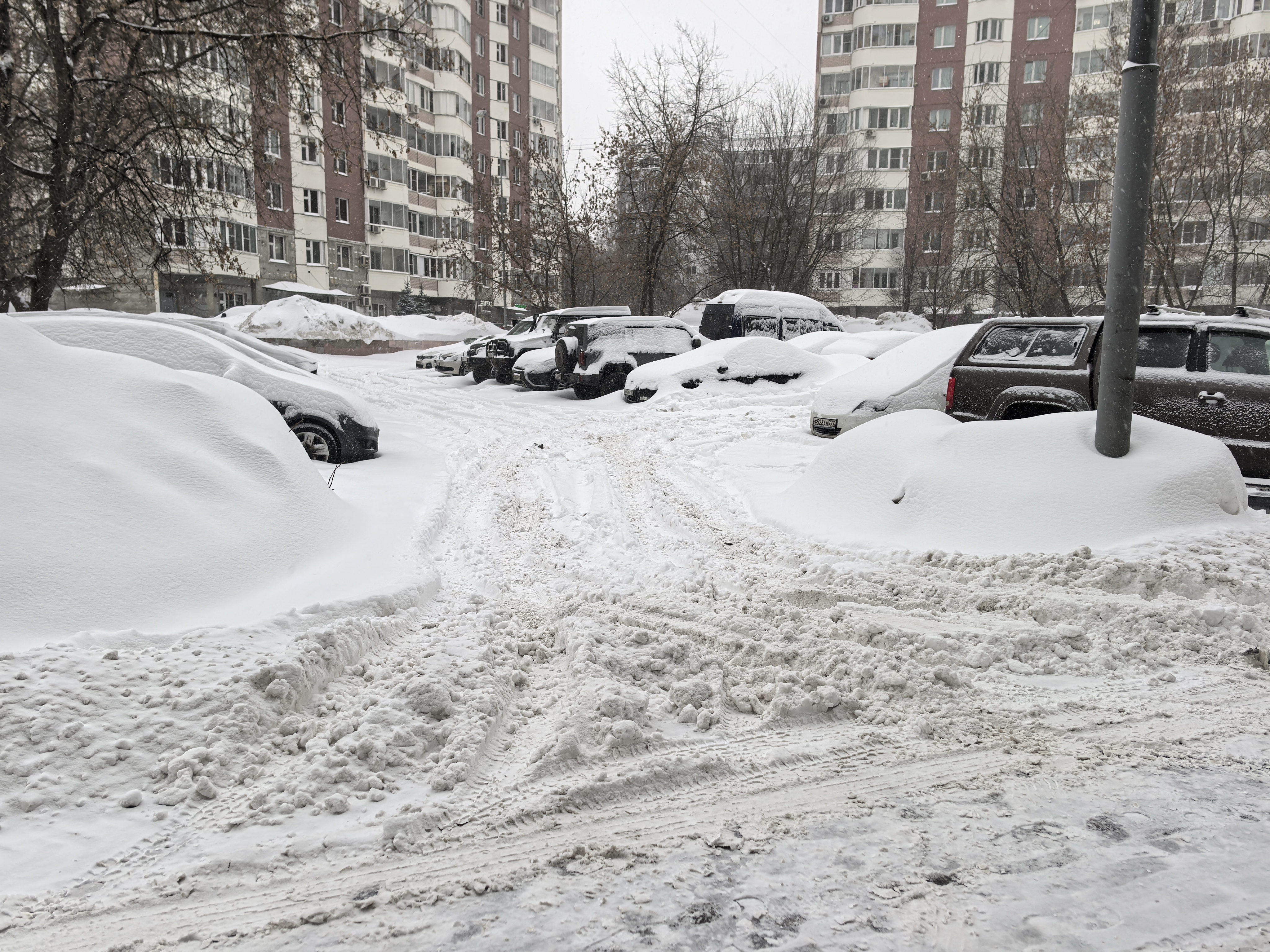 Дворовая территория с парковкой между жилыми домами, полностью покрытая снегом. Проезжая часть узкая, образованная следами от шин, припаркованные автомобили занимают значительную часть пространства.