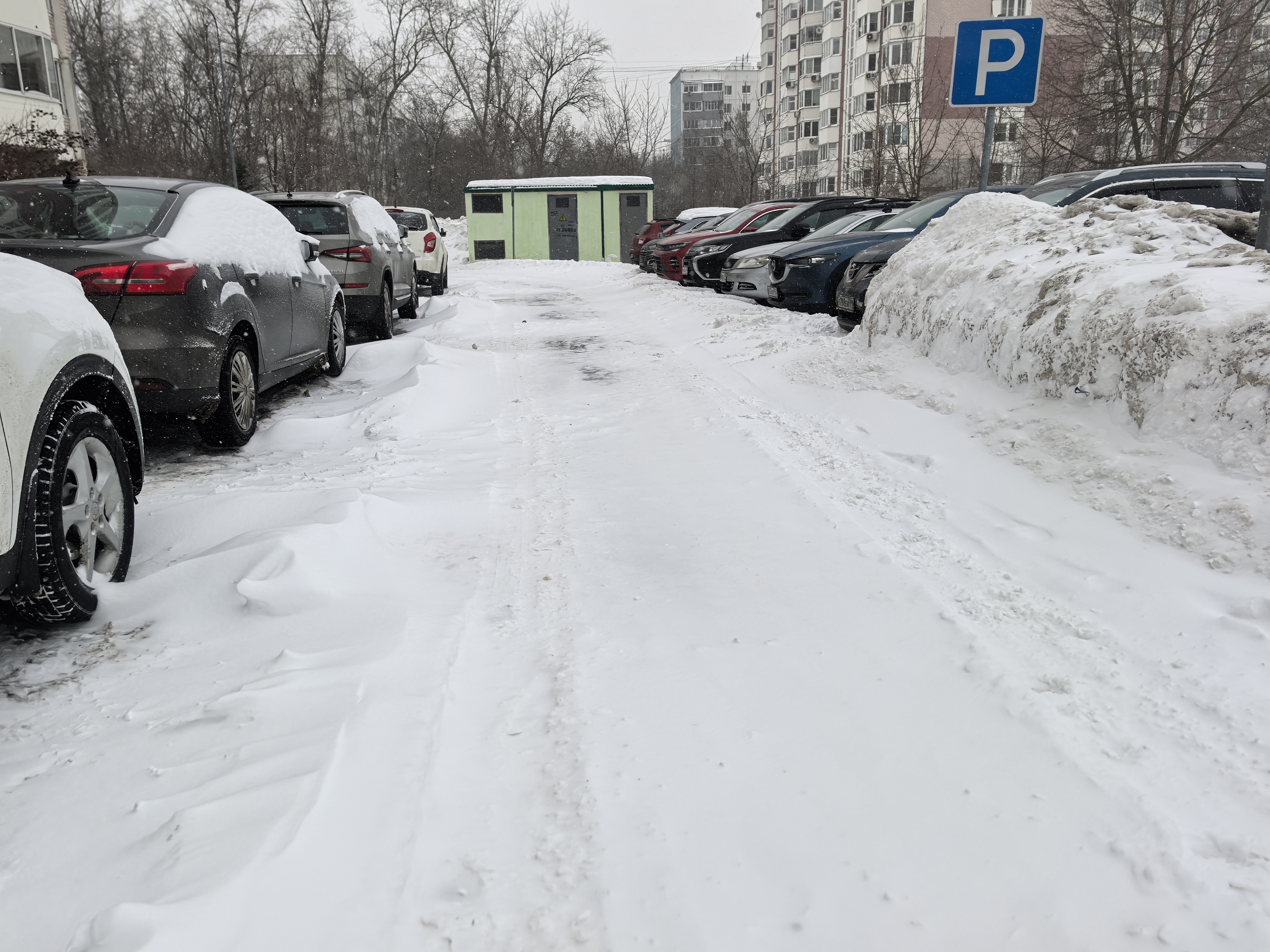Дворовая территория с парковкой, покрытая снегом. Видны припаркованные автомобили с обеих сторон проезжей части, жилые дома на заднем плане. Переходов и тротуаров не видно.