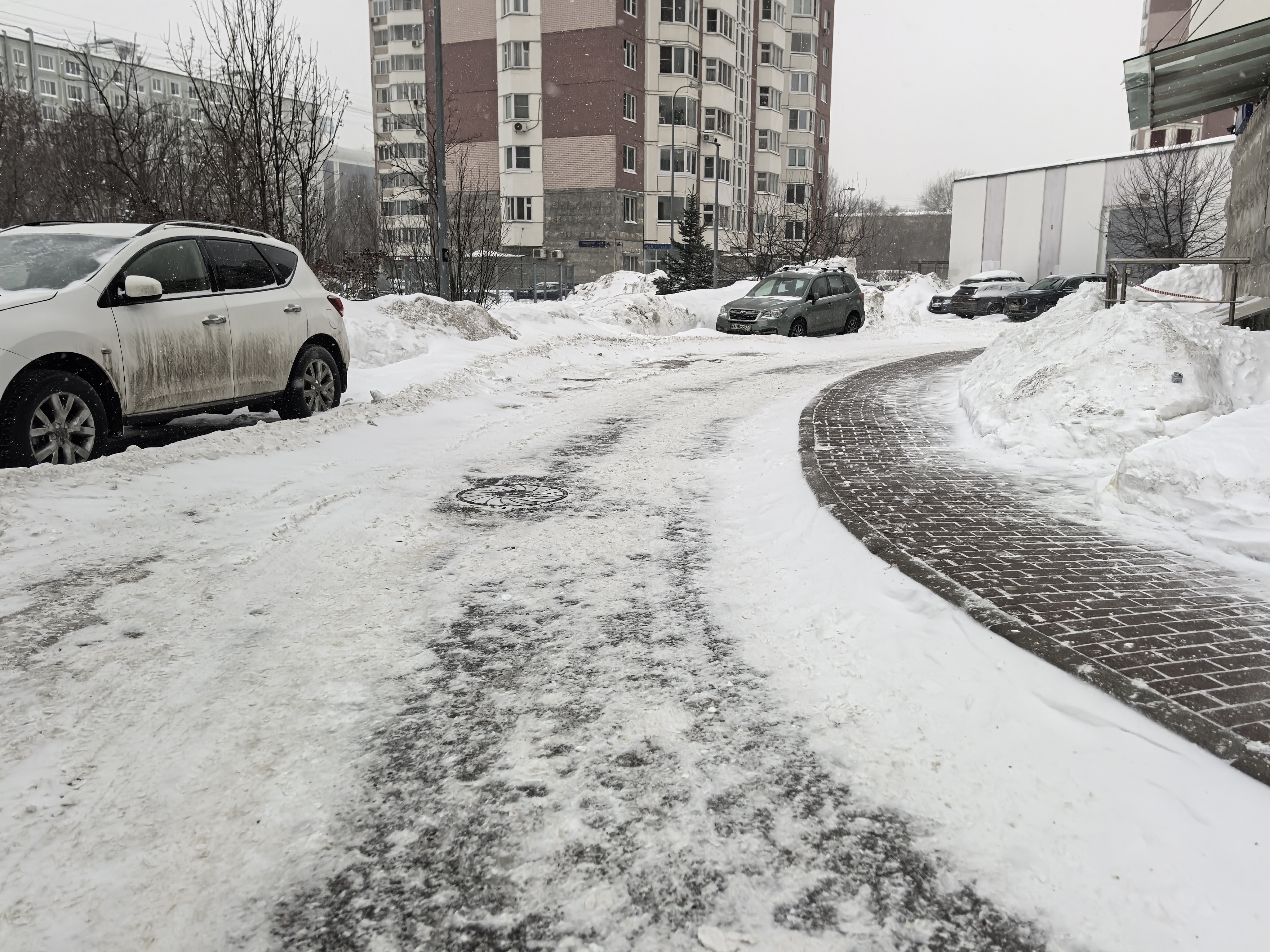 Дворовая территория с проезжей частью, покрытой снегом и льдом, и тротуаром из брусчатки. Видны припаркованные автомобили и жилые дома.