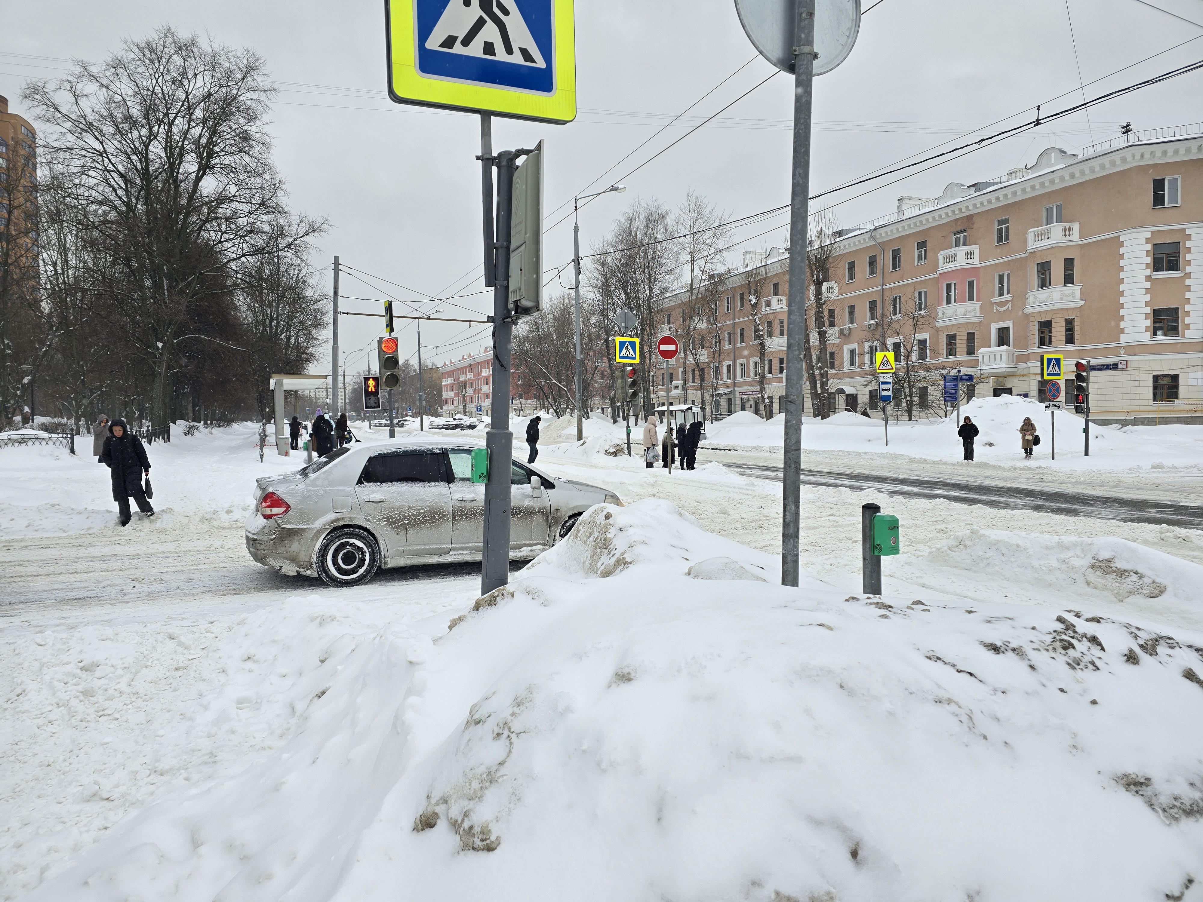 Перекресток с пешеходным переходом, регулируемым светофором. Зимние условия, много снега на обочинах и тротуарах.