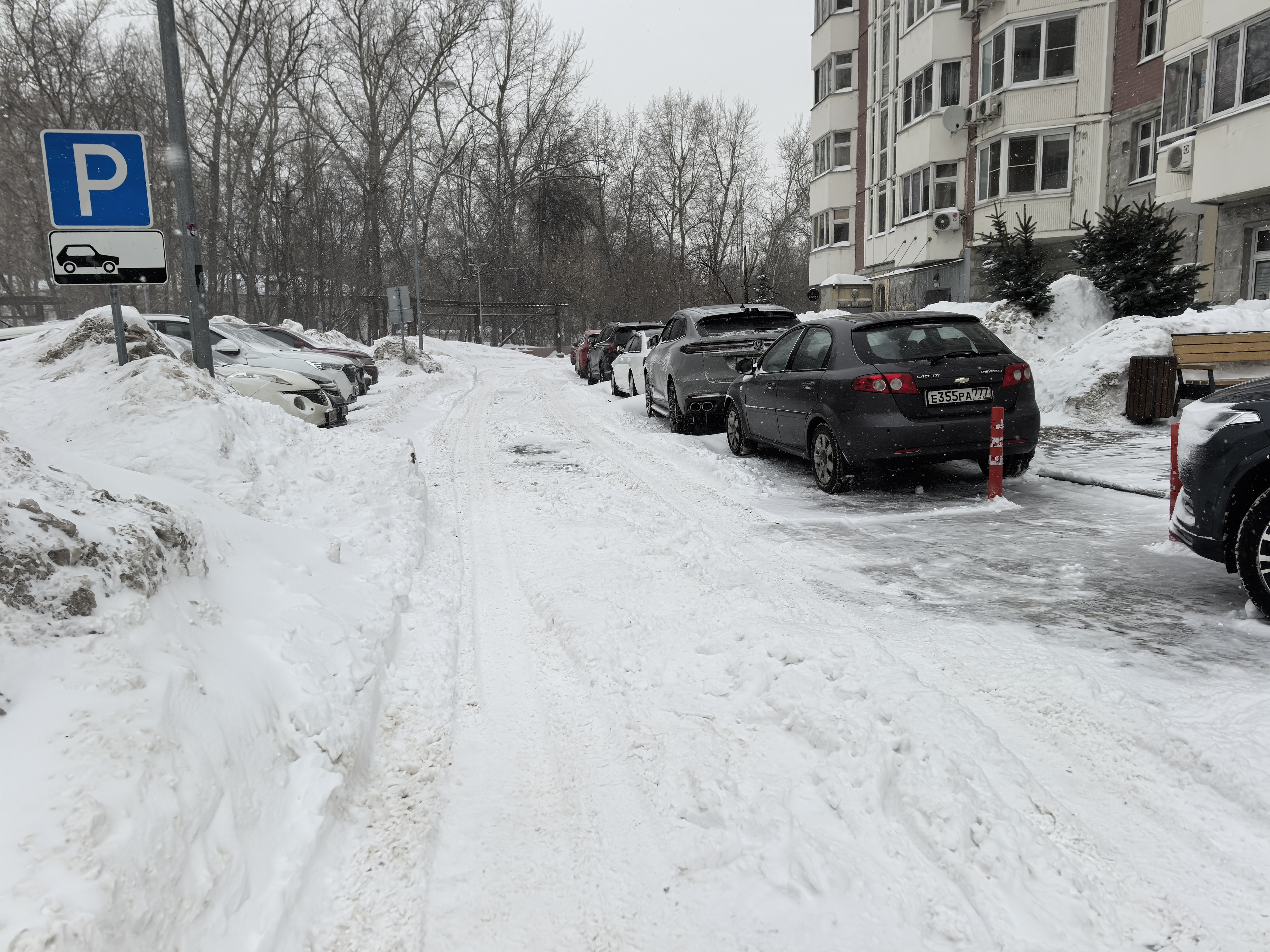 Дворовая территория с припаркованными автомобилями, покрытая снегом и льдом. Видны жилые дома, деревья и знаки парковки. Дорога не имеет разметки и пешеходных переходов, используется как проезд и парковка.