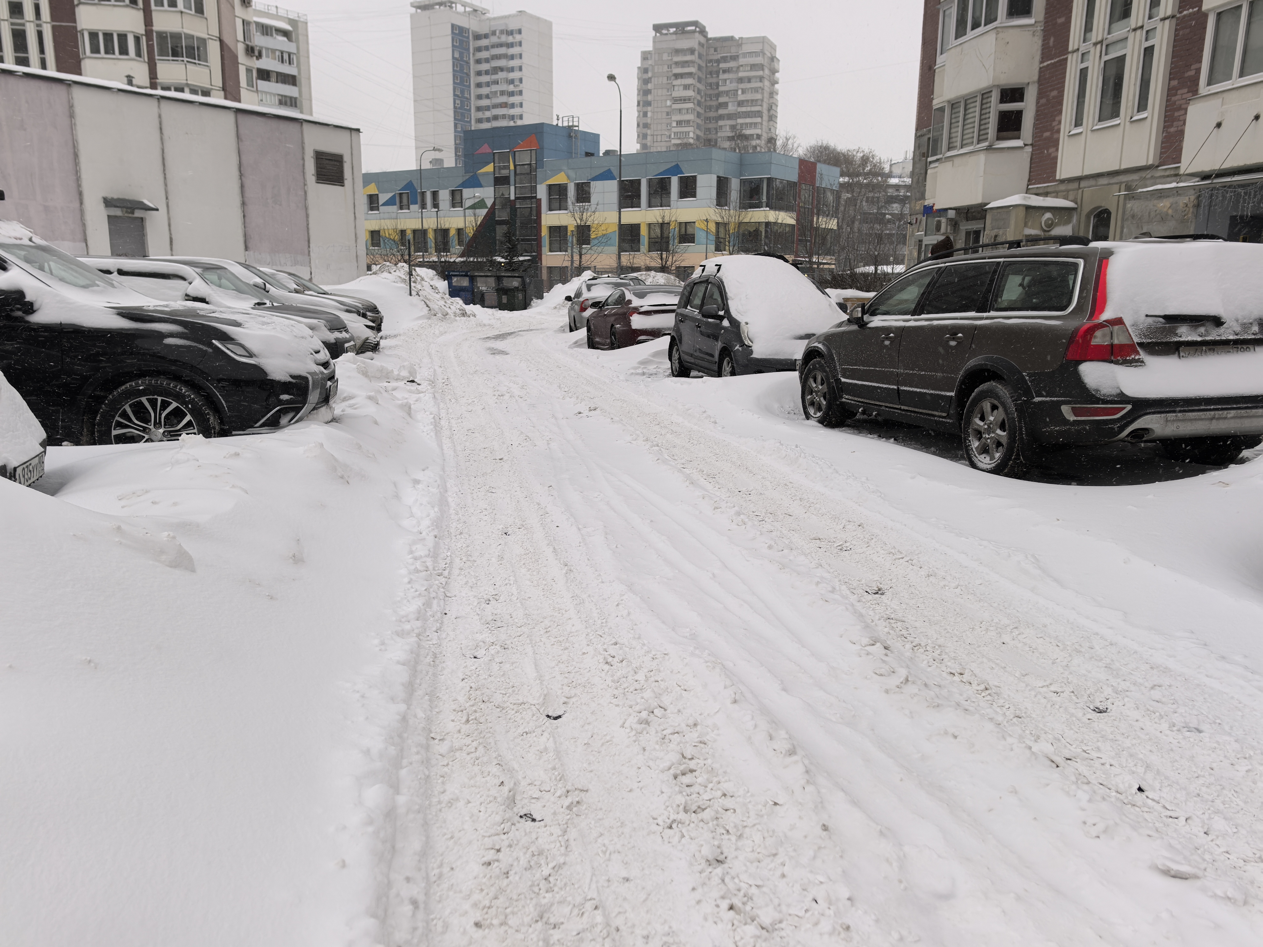 Дворовая территория с парковкой автомобилей, покрытая снегом. На заднем плане виден детский сад или школа.