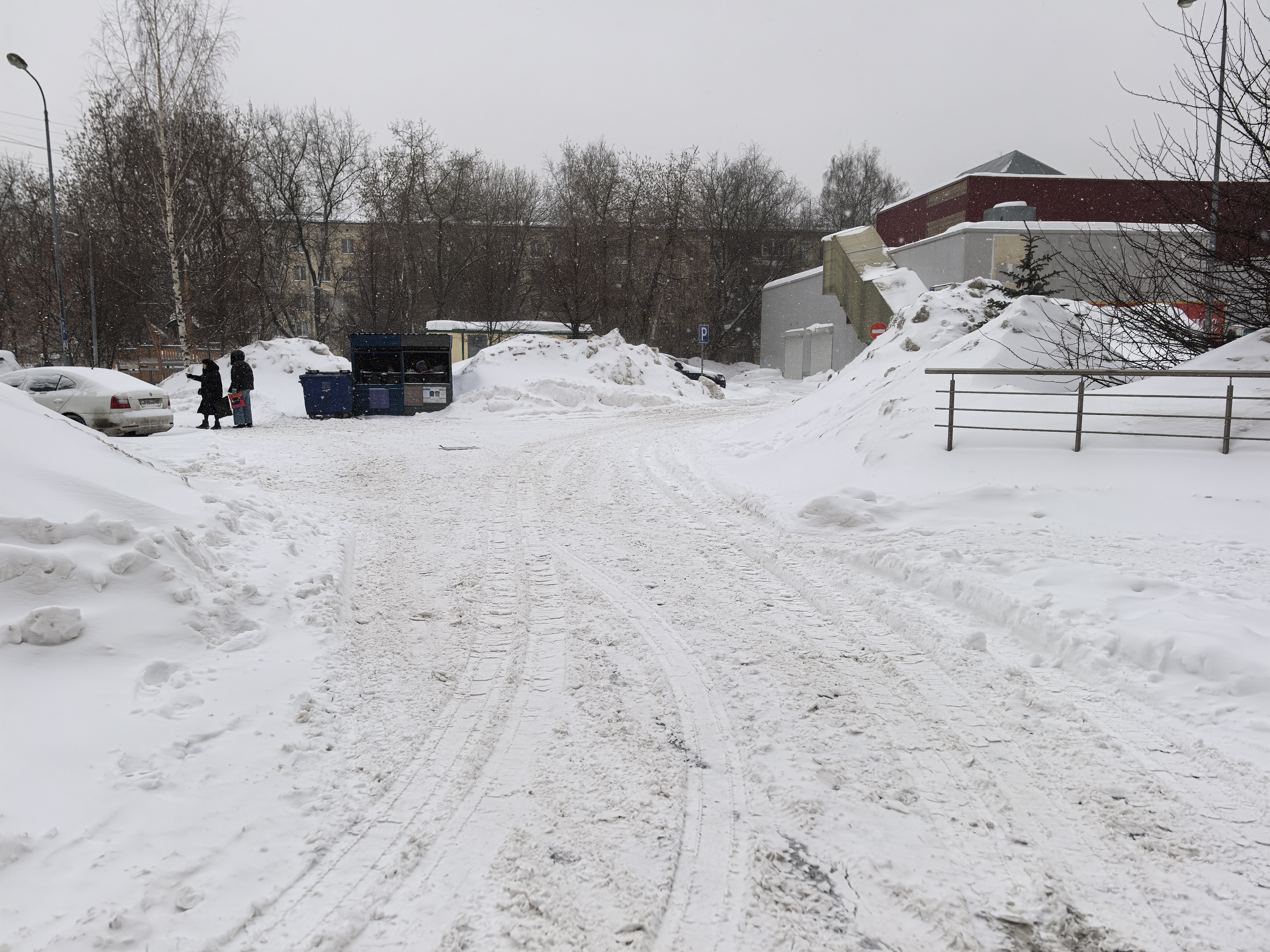 Дворовая территория, заснеженная дорога с колеями от машин, контейнеры для мусора, припаркованный автомобиль и дети на обочине.