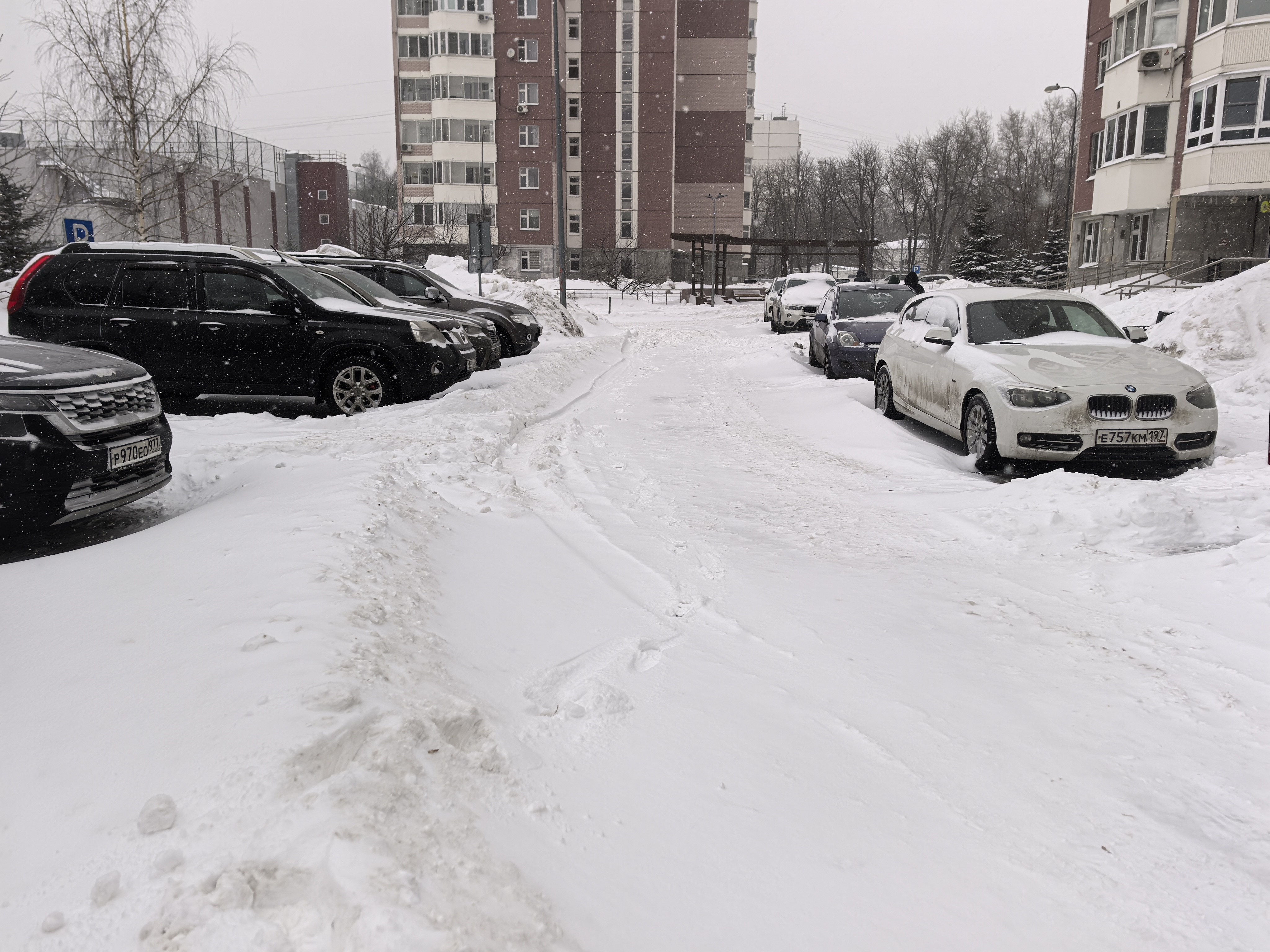 Дворовая территория, покрытая снегом, с припаркованными автомобилями по обеим сторонам проезжей части. Видны жилые дома и ограждения.