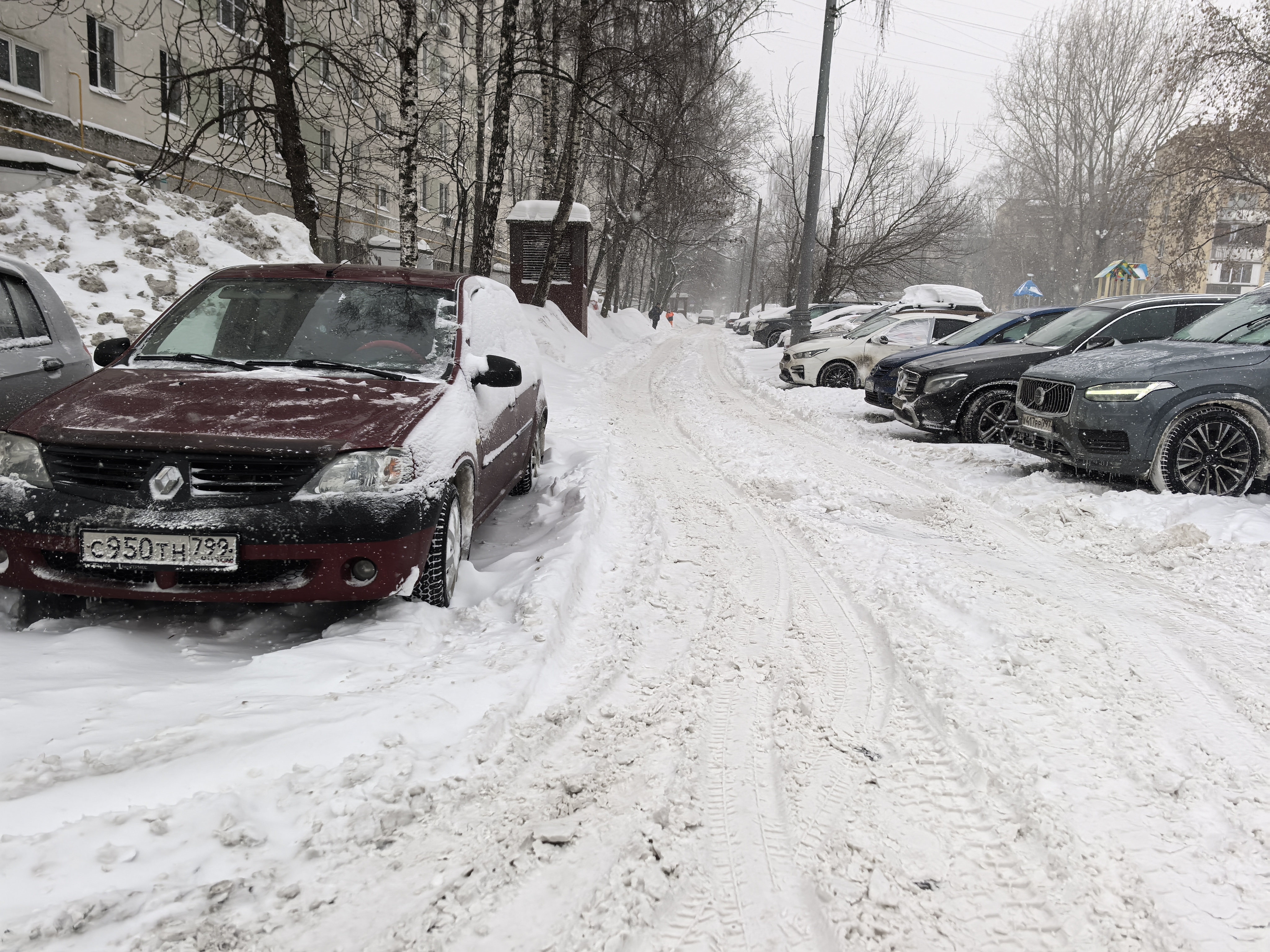 Дворовая территория, покрытая снегом, с припаркованными автомобилями по обеим сторонам проезжей части. Вдали видна детская площадка.