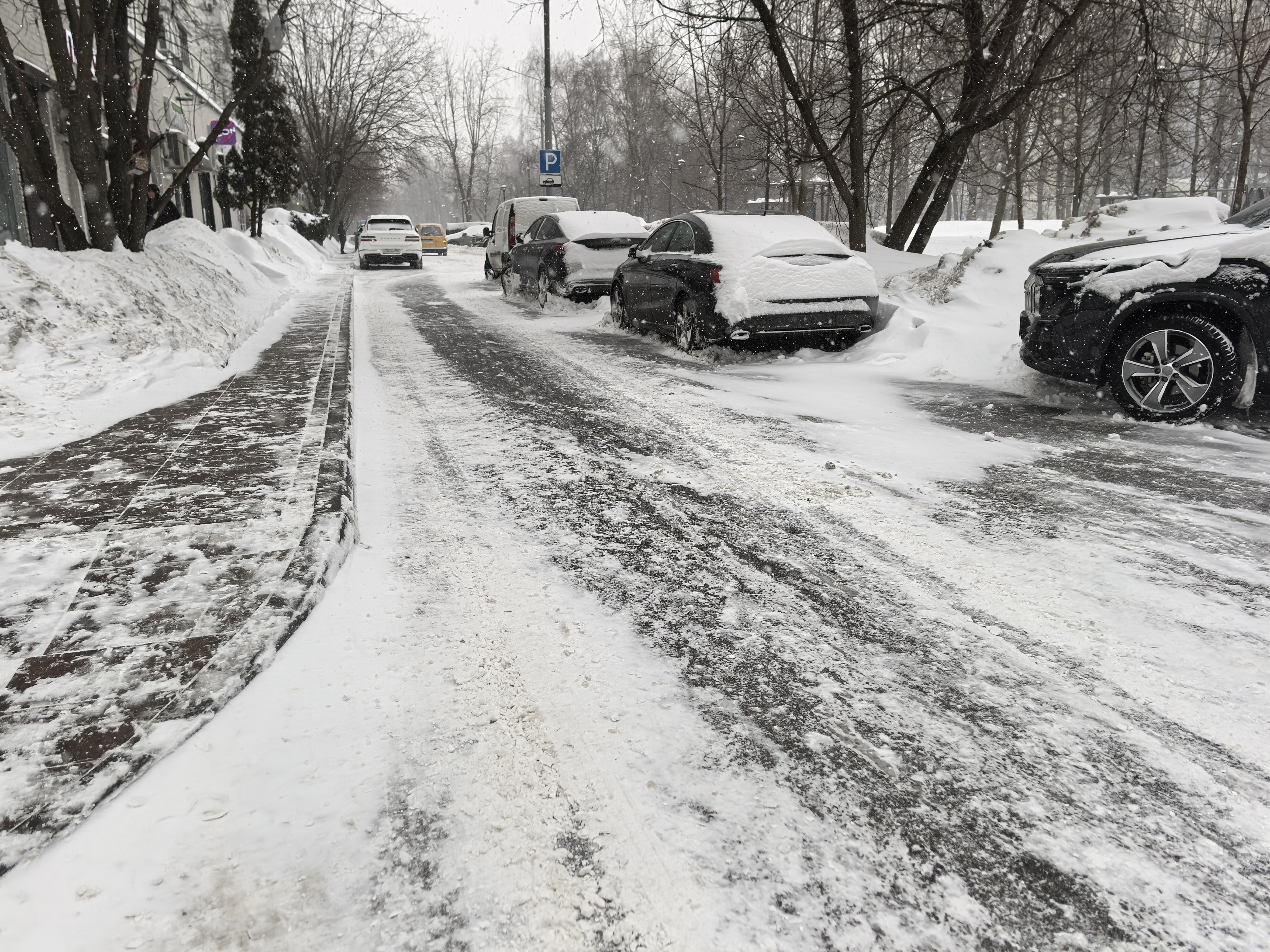 Участок городской улицы в зимний период. Видна проезжая часть, покрытая снегом и слякотью, с припаркованными автомобилями. Слева — тротуар, частично очищенный от снега. Пешеходный переход на видимом участке отсутствует.