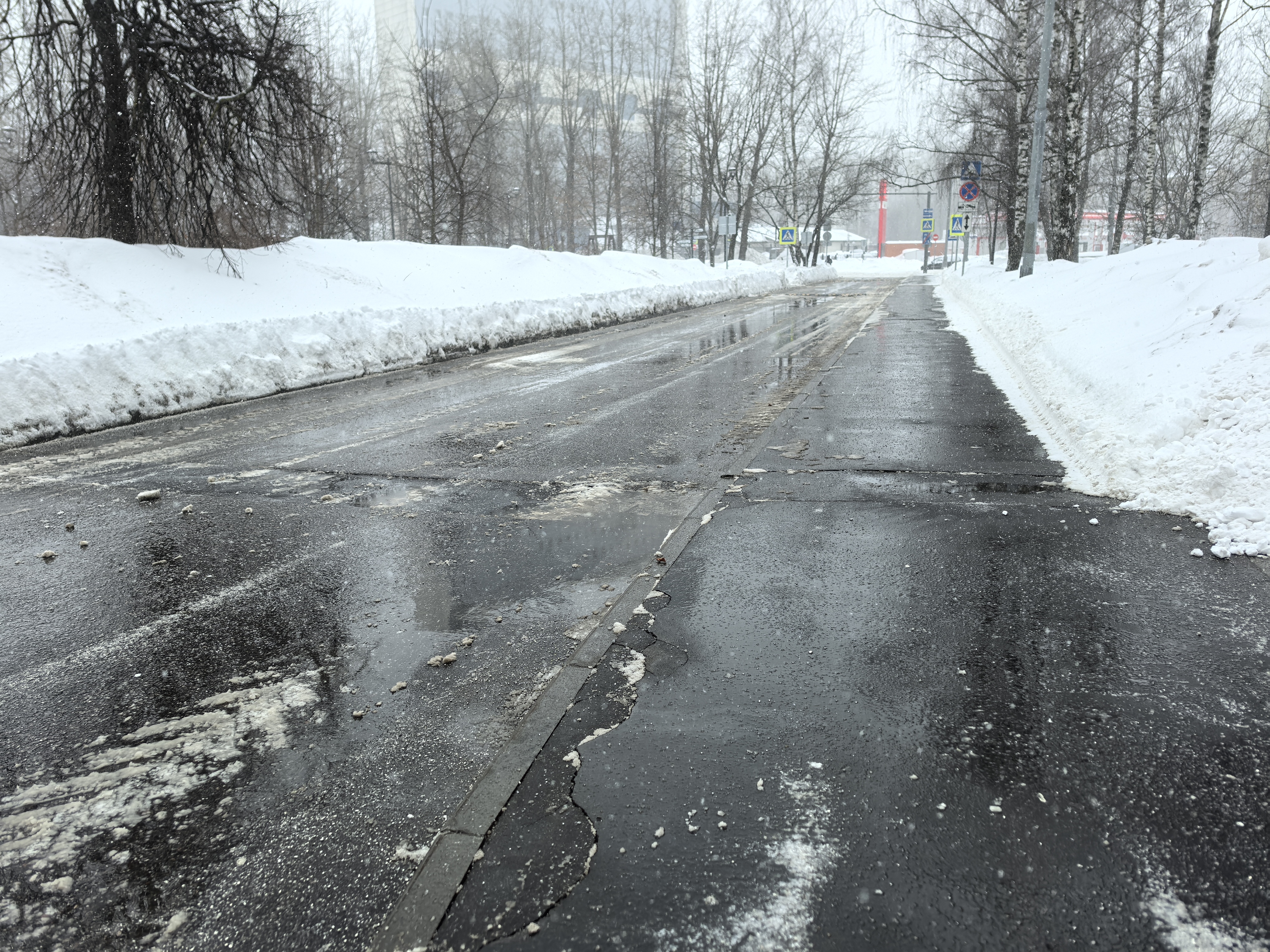 Проезжая часть с пешеходным переходом в зимних условиях. На дороге видна мокрая поверхность, ледяная корка и остатки снега. Вдали видны знаки пешеходного перехода, но сама зебра не просматривается из-за погодных условий.