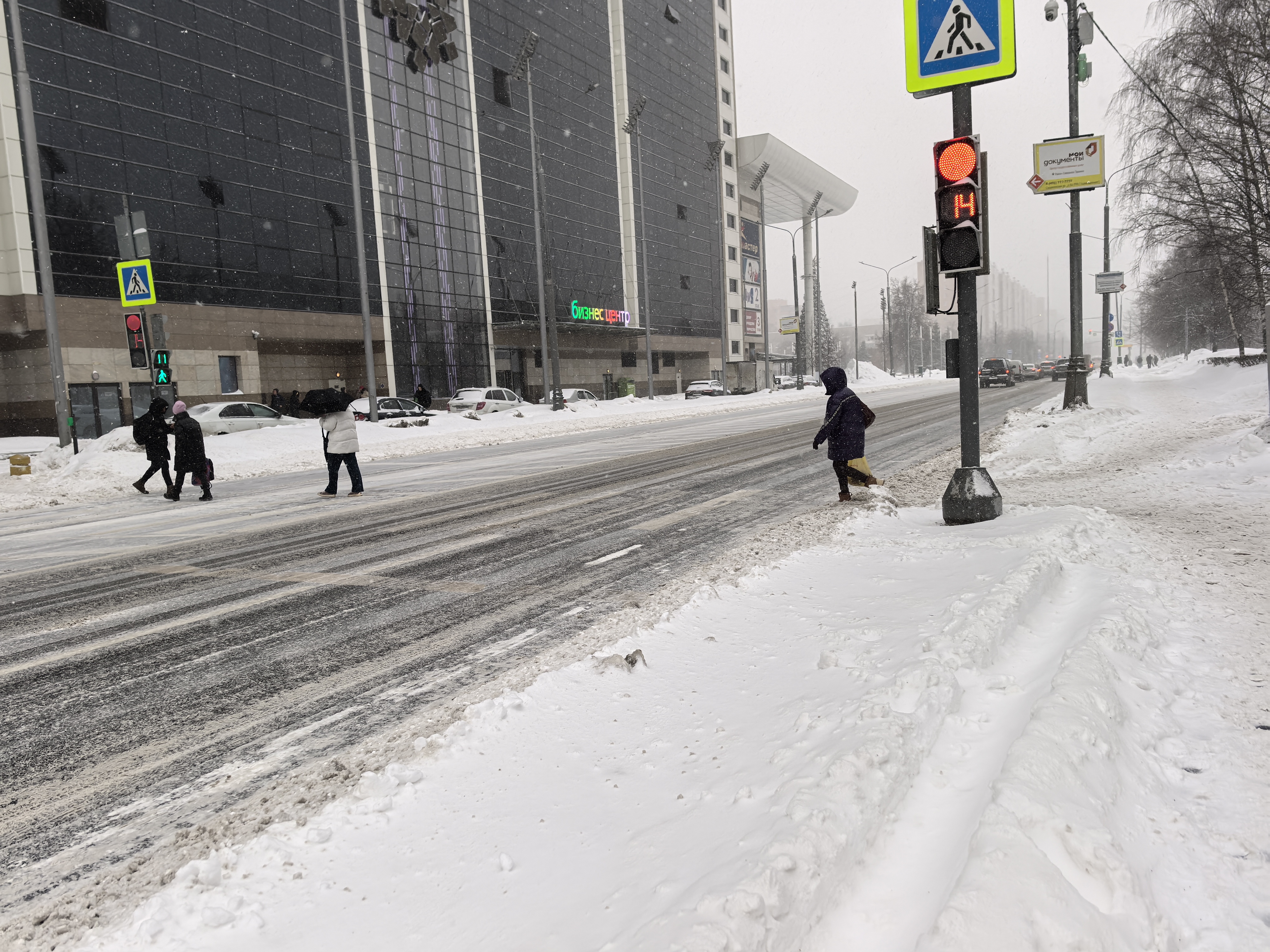 Городская улица с пешеходным переходом, регулируемым светофором, в зимних условиях (снег, гололед). Видны пешеходы, переходящие дорогу.