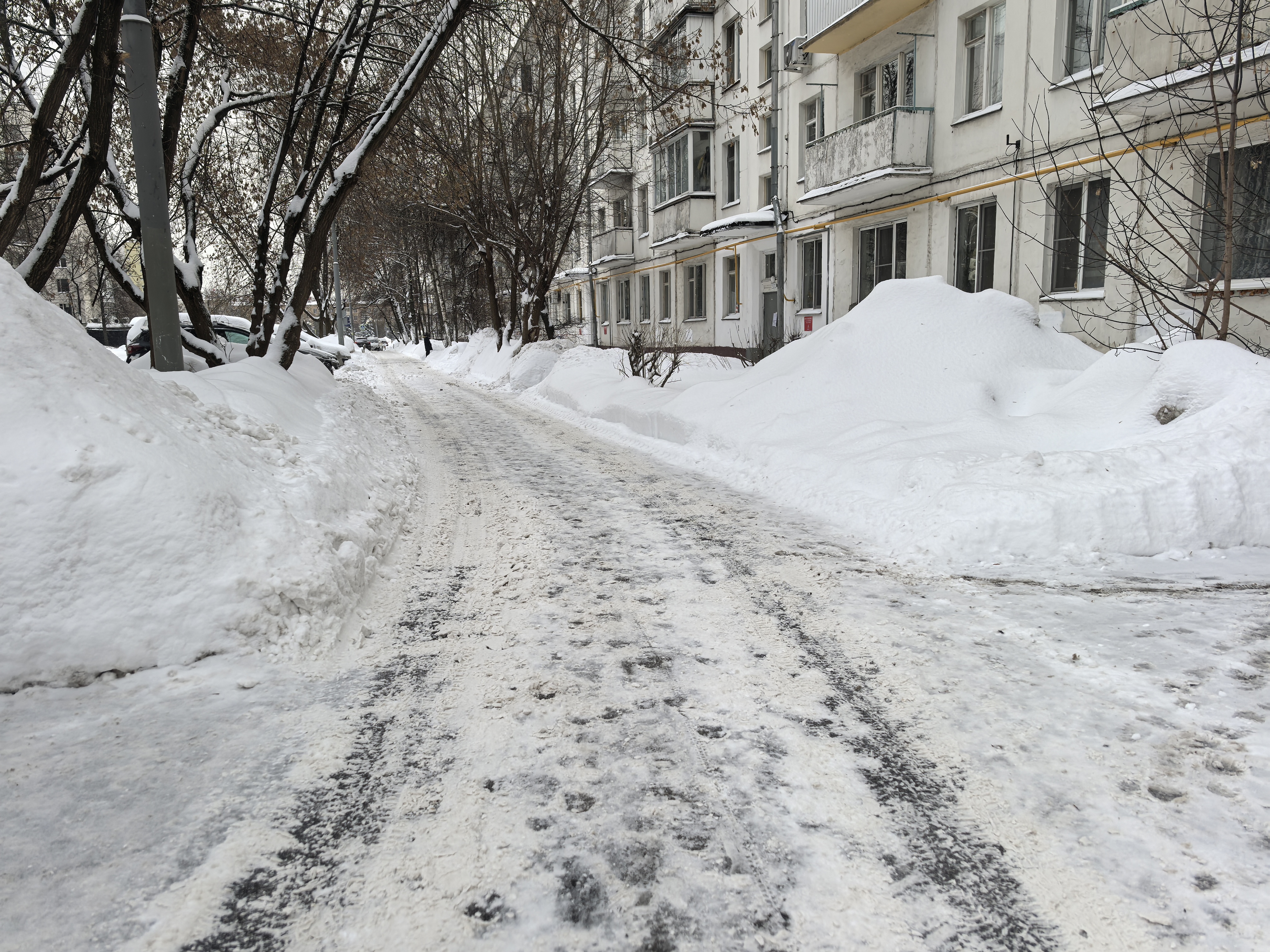 Дворовая территория между жилыми домами, покрытая снегом и льдом. Тротуары полностью засыпаны снегом, проезжая часть узкая, с видимыми следами от шин и пешеходов.